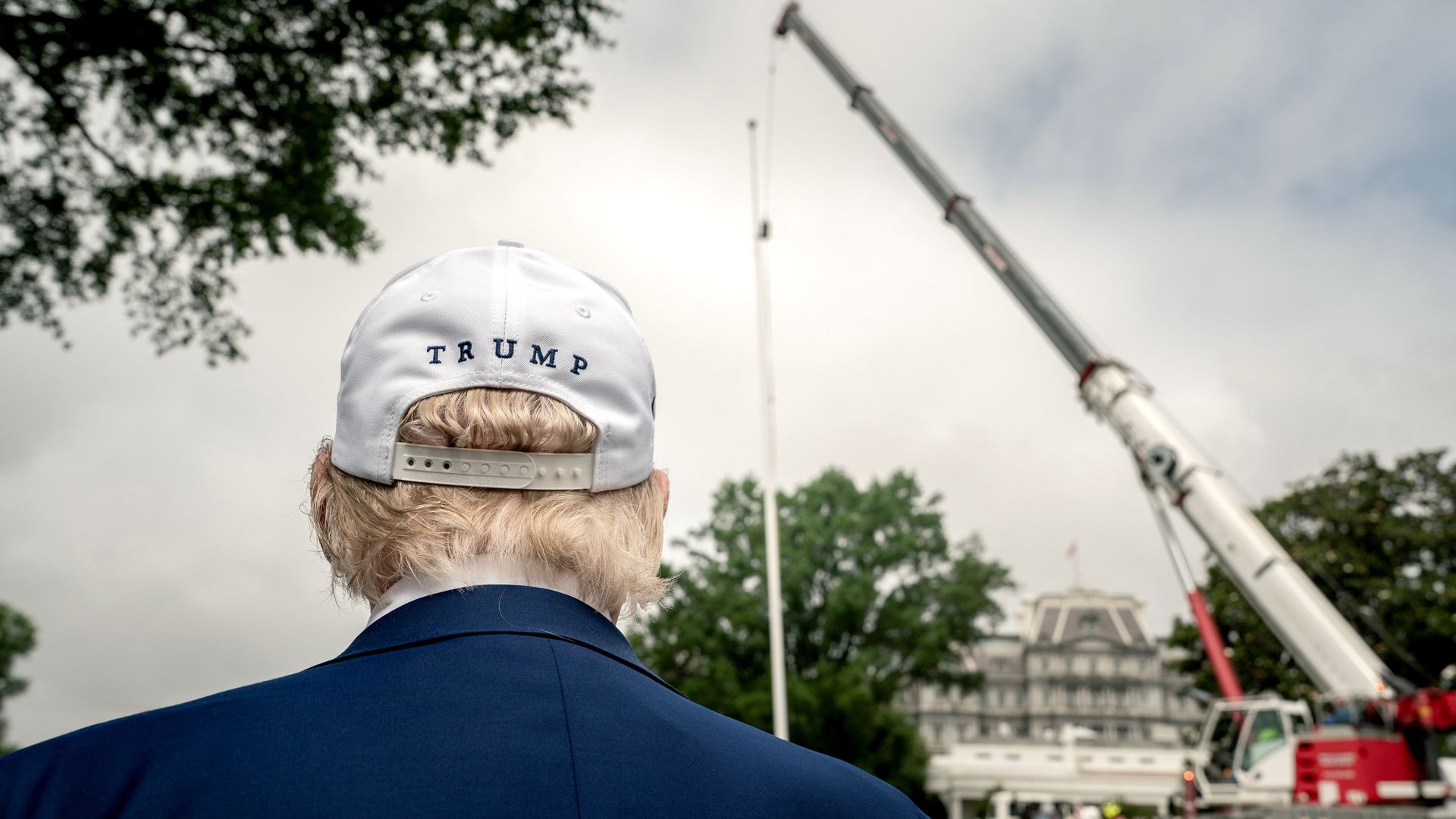 A photograph of Donald Trump facing away from the camera, watching a crane lift a flag pole