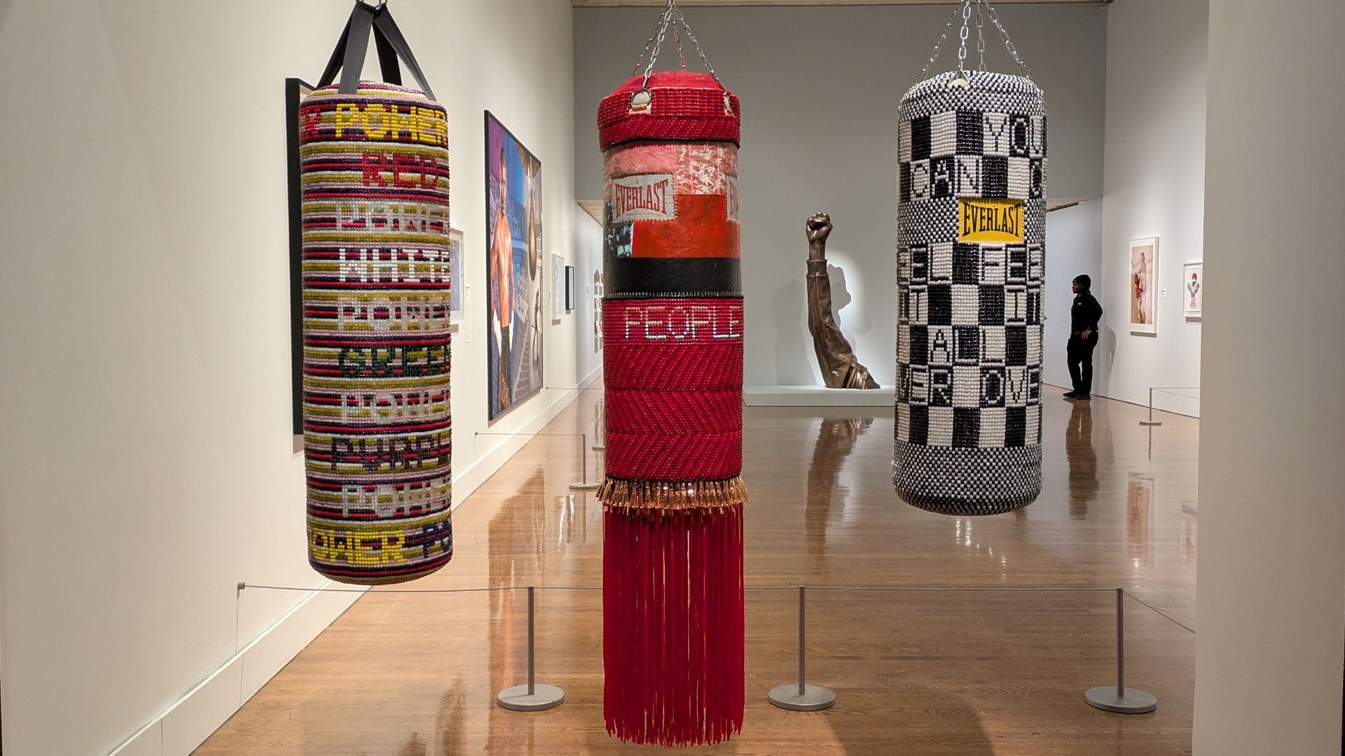 Museum gallery with three tall hanging cylindrical sculptures: left colorful stripes, middle red/black labeled EVERLAST and PEOPLE, right black-and-white checkerboard. A man stands in the background.