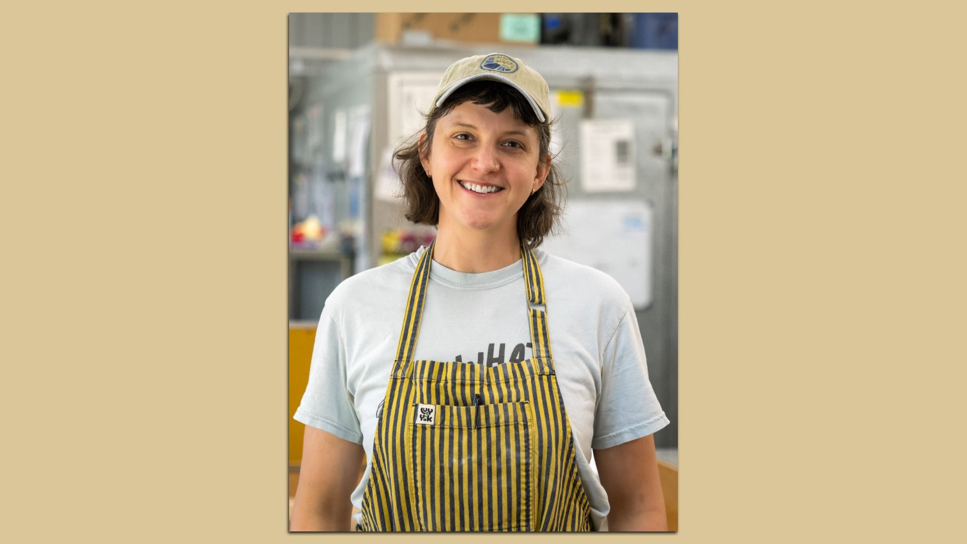 Portrait of a smiling woman in a beige cap and striped yellow-and-navy apron in a busy kitchen, with stainless steel equipment in the background.