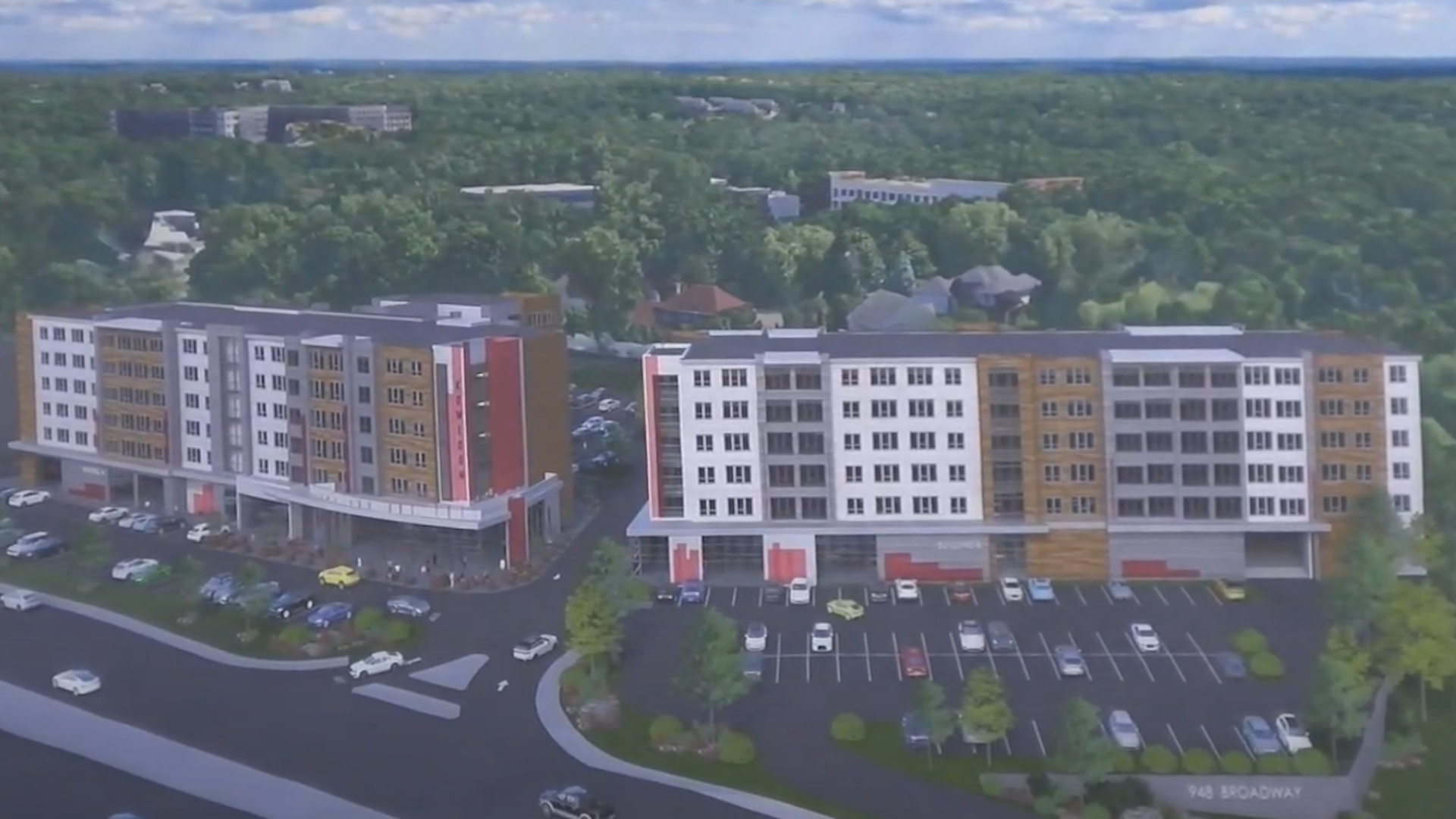 Aerial view of two large, modern six-story apartment buildings with white and brown paneling, surrounded by parking lots filled with cars and green trees under a partly cloudy sky.