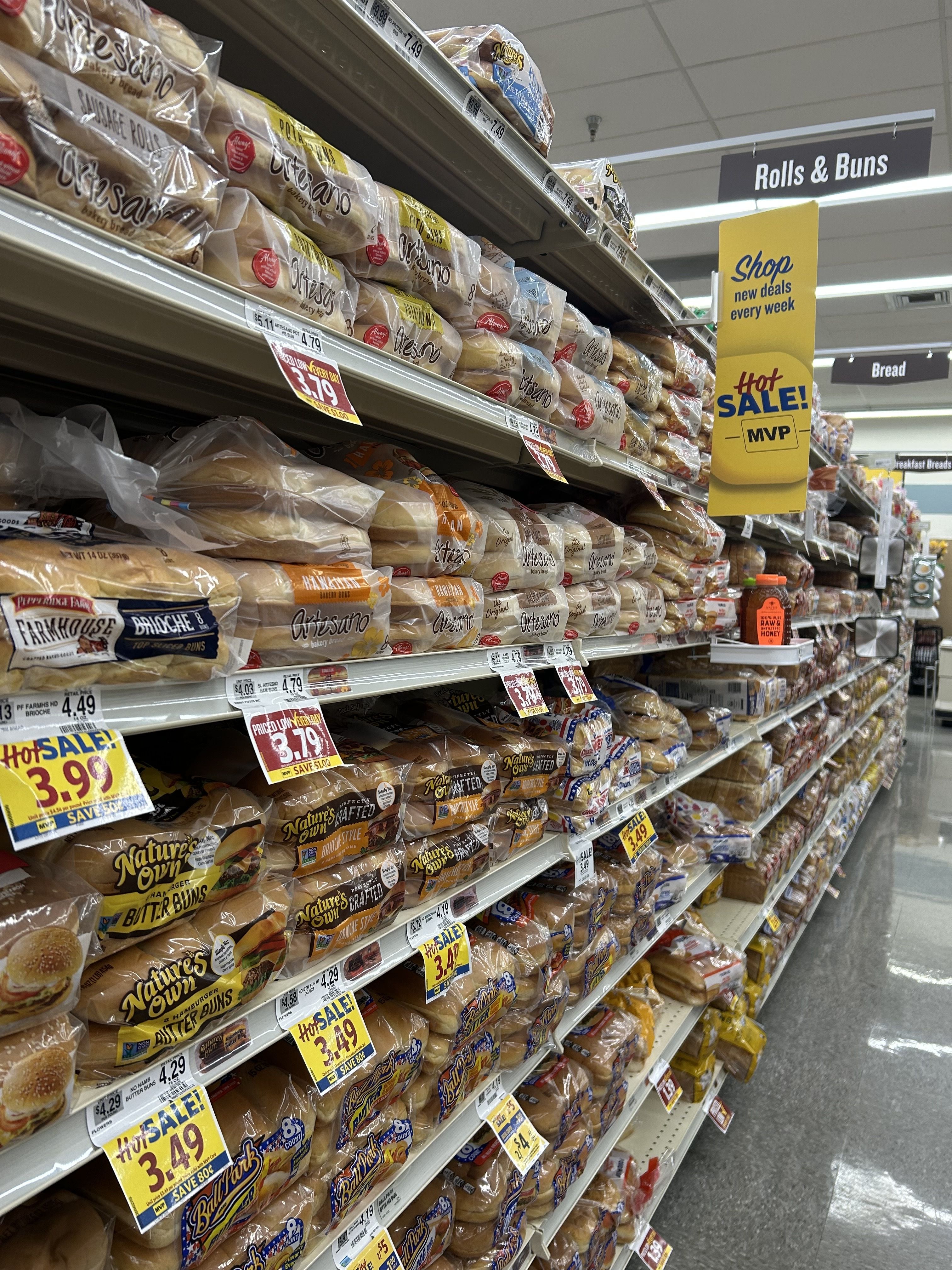 Supermarket bread aisle with shelves of bagged sandwich rolls and buns, bright yellow sale signs, and an overhead sign reading Rolls & Buns.