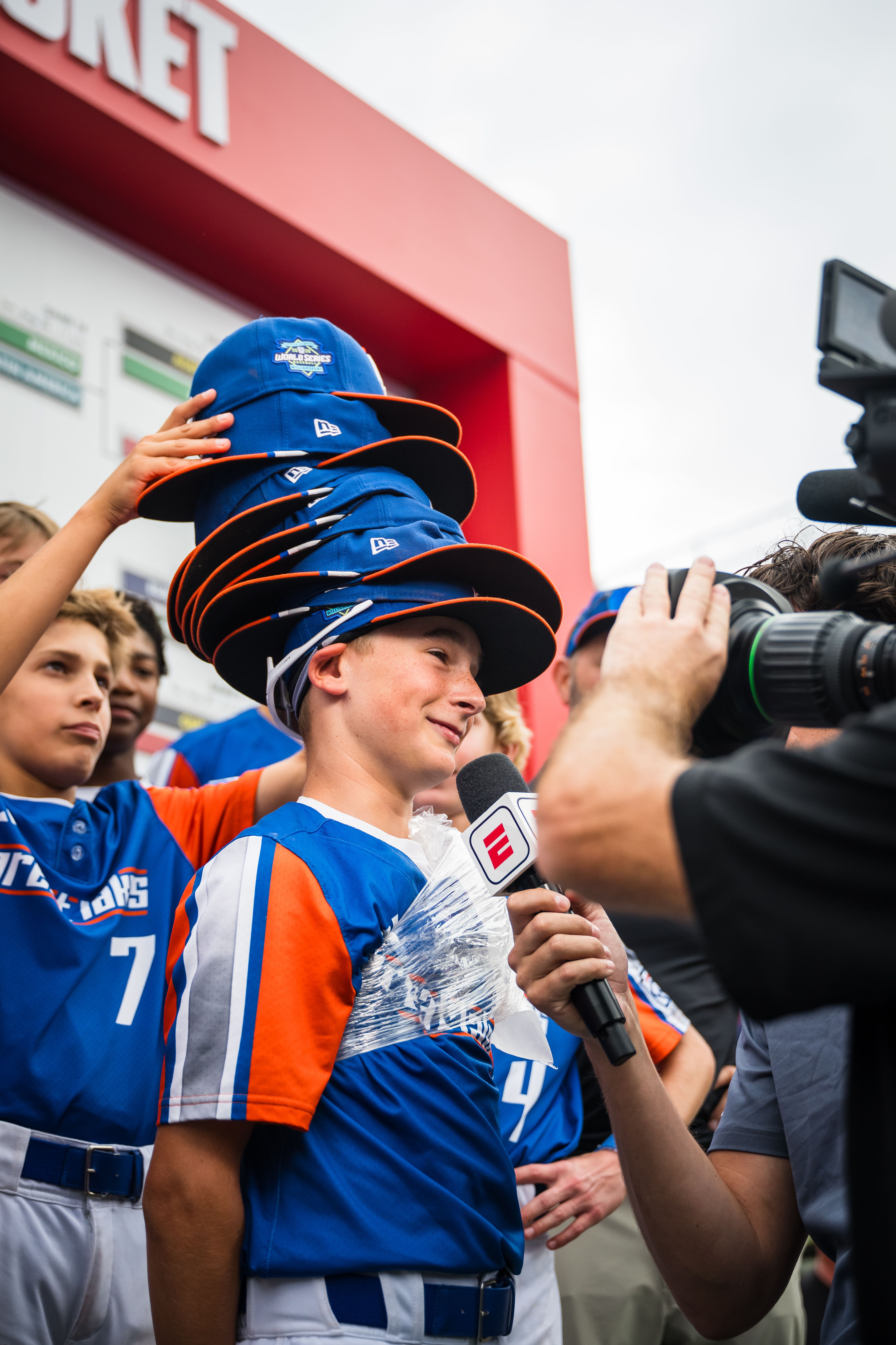 Young man in a blue and orange jersey balances a towering stack of blue caps with orange brims on his head, while a reporter with an ESPN microphone and a camera crew film nearby near a red backdrop.