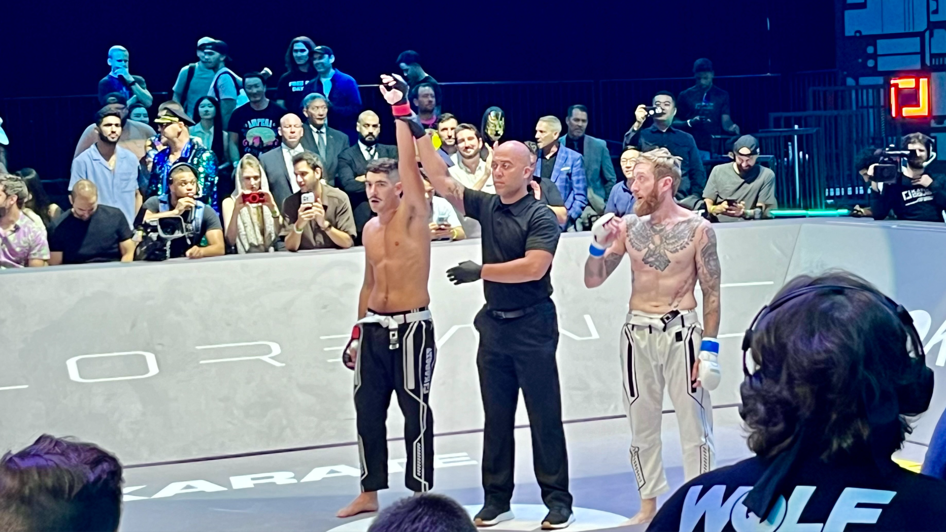 Two men in a ring at the end of a fight, with an official indicating one as winner. While spectators look on.