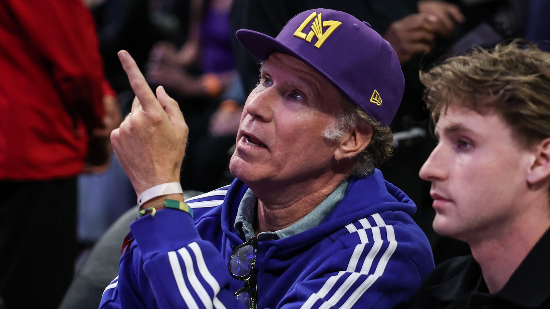 A man in a purple cap with a yellow emblem and a purple jacket with white stripes gestures upward with his index finger; a younger man sits to his right in a crowded setting.