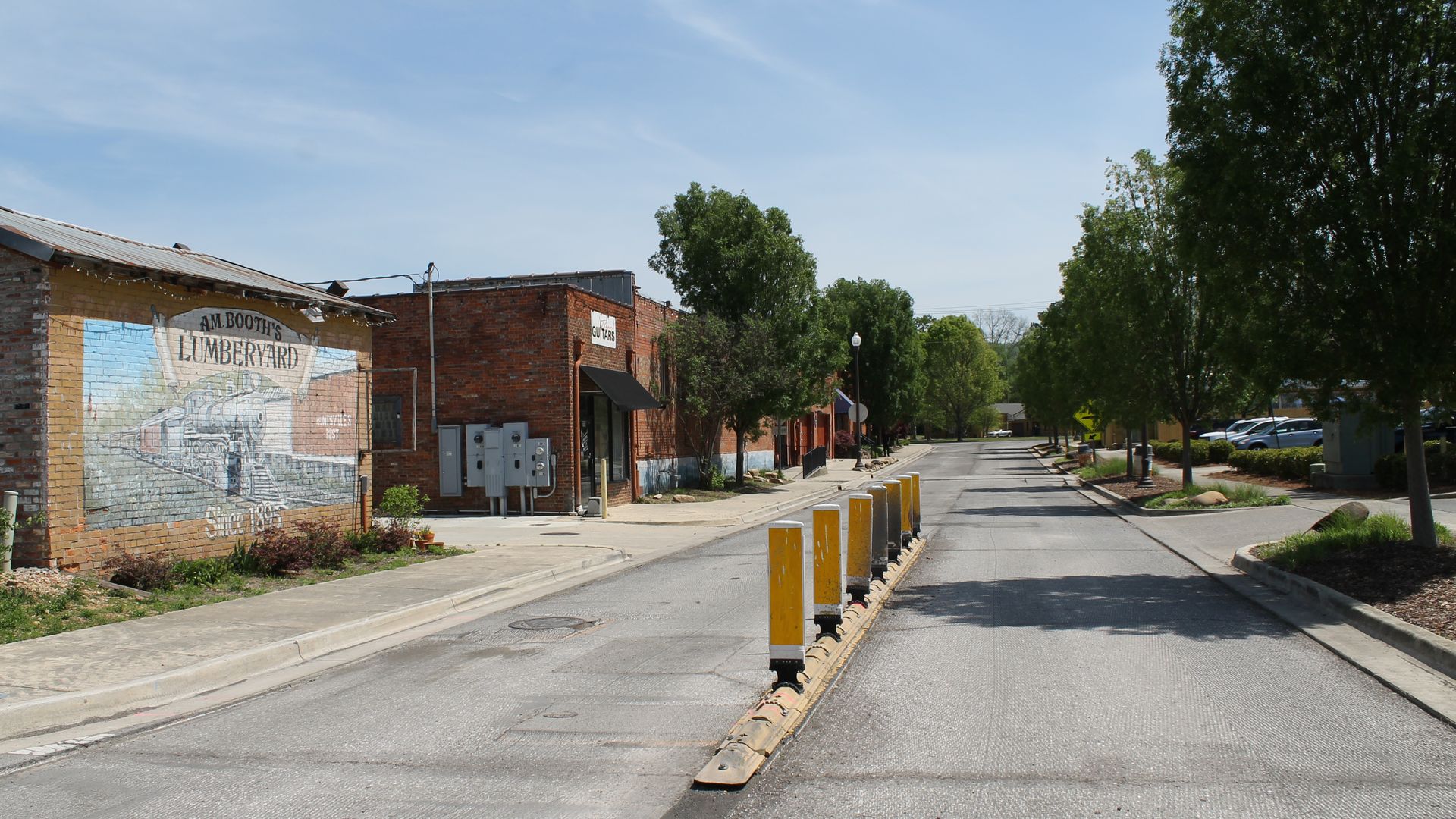 Sunny small-town street with brick buildings and a mural reading "AM Booth's Lumberyard" on the left. A row of yellow bollards runs down a center barrier, with trees along the sidewalk and distant cars.