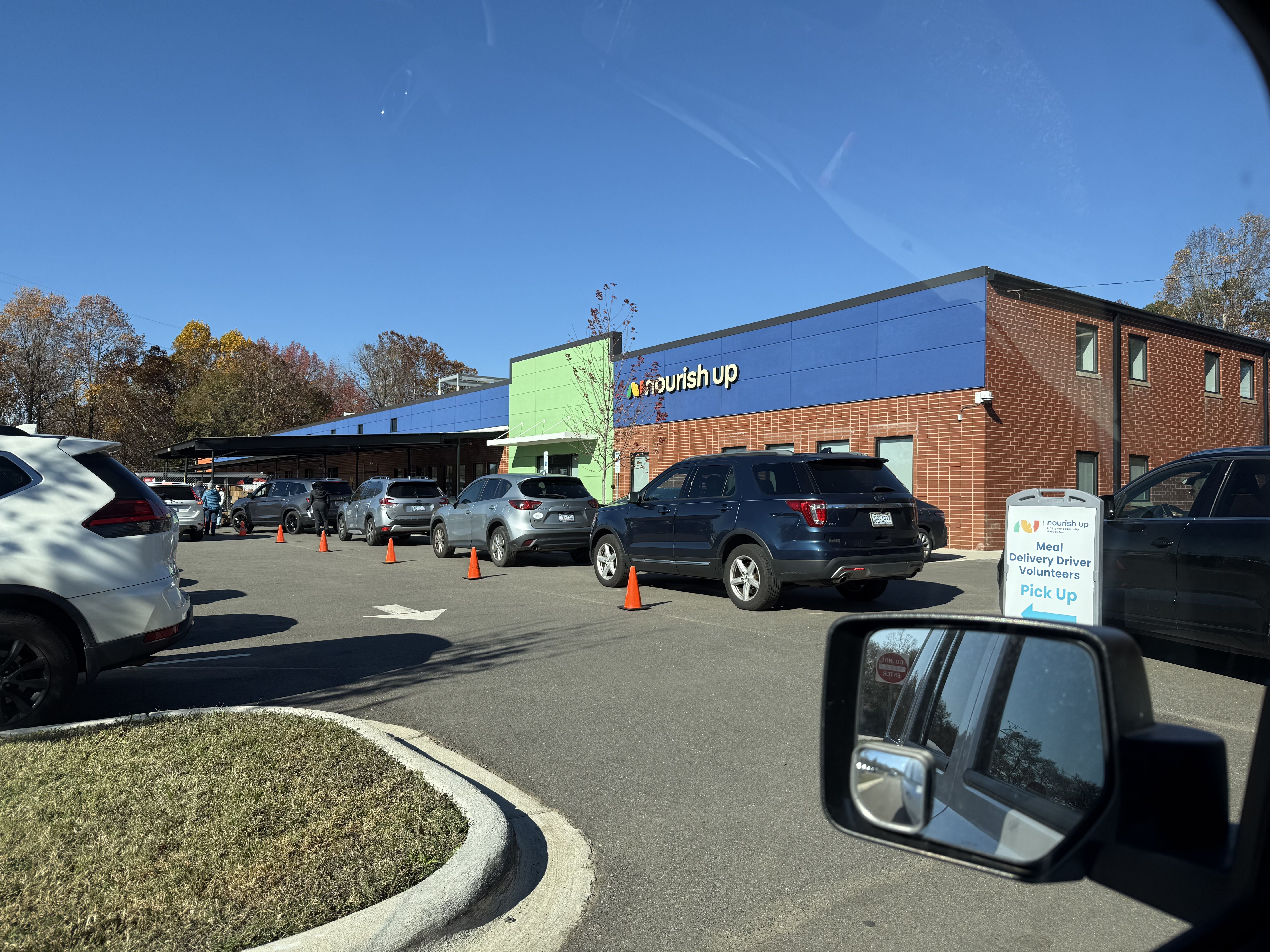 Line of cars and volunteers outside a brick building with blue and green accents at "nourish up" for Meal Delivery Driver Volunteer Pick Up on a clear, sunny day.