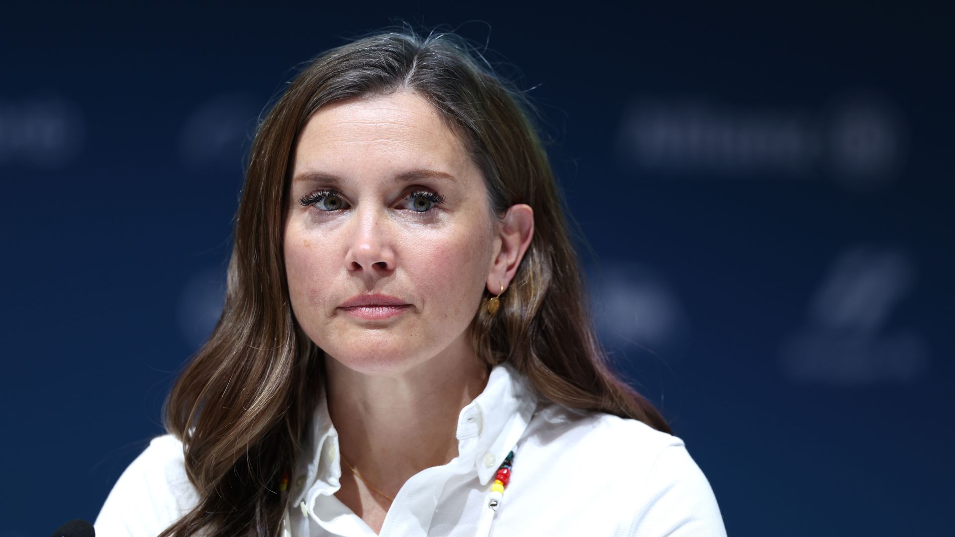 A woman with long brown hair, wearing a white shirt and a colorful beaded necklace, sits at a panel with a microphone, against a dark blue backdrop.