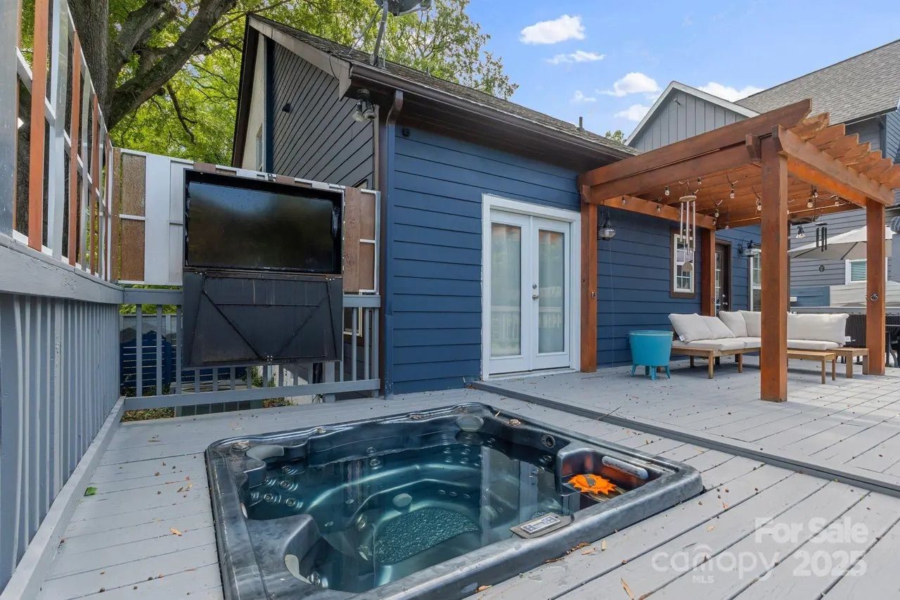 Outdoor deck with built-in hot tub, blue house exterior, white French doors, wooden pergola with string lights, and cushioned seating under the pergola on a clear day.