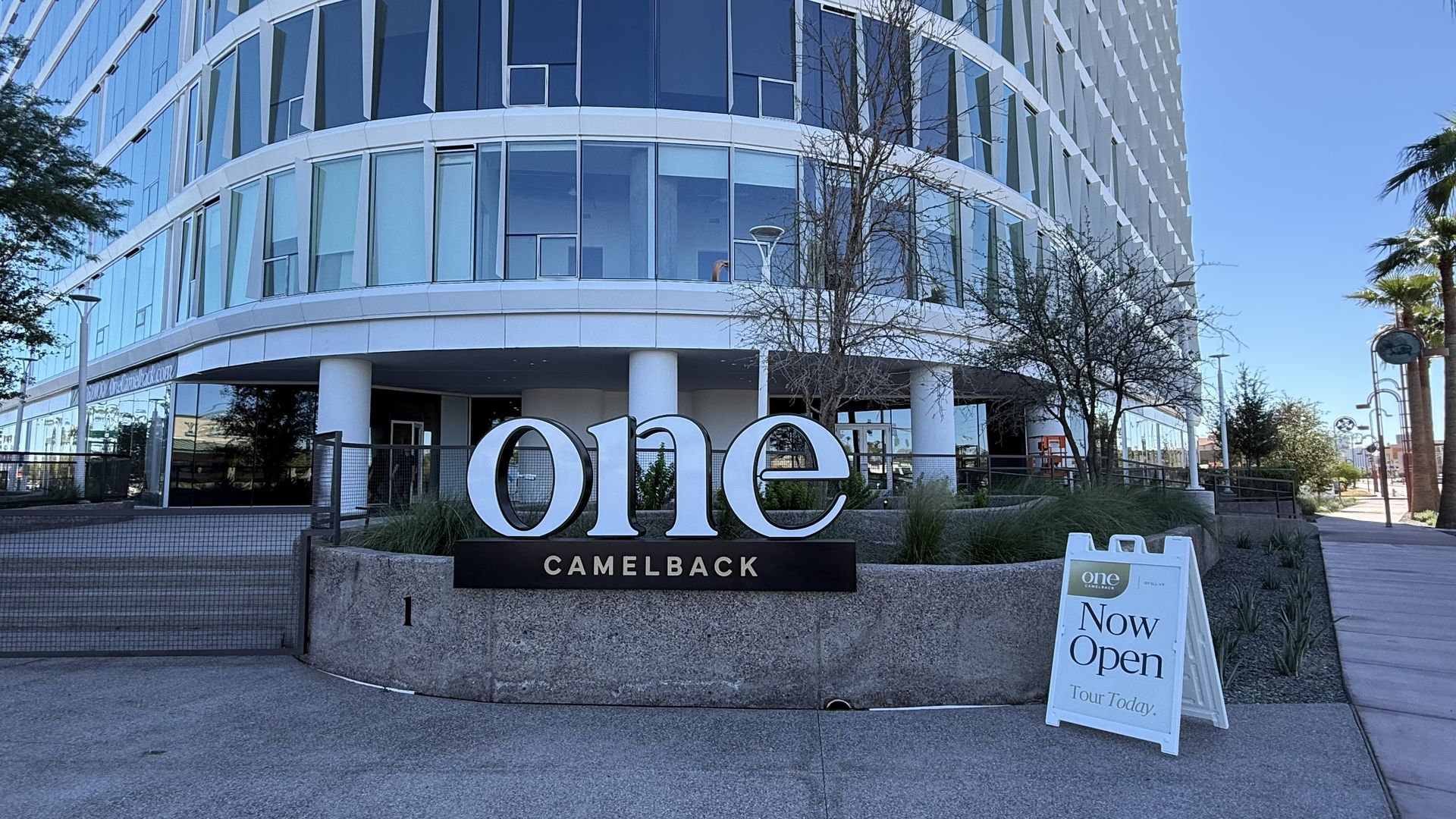 Modern curved glass office-style building with white framing and a large ONE sign; Camelback below. A 'Now Open' sidewalk sign sits by leafless trees and palms against a blue sky.