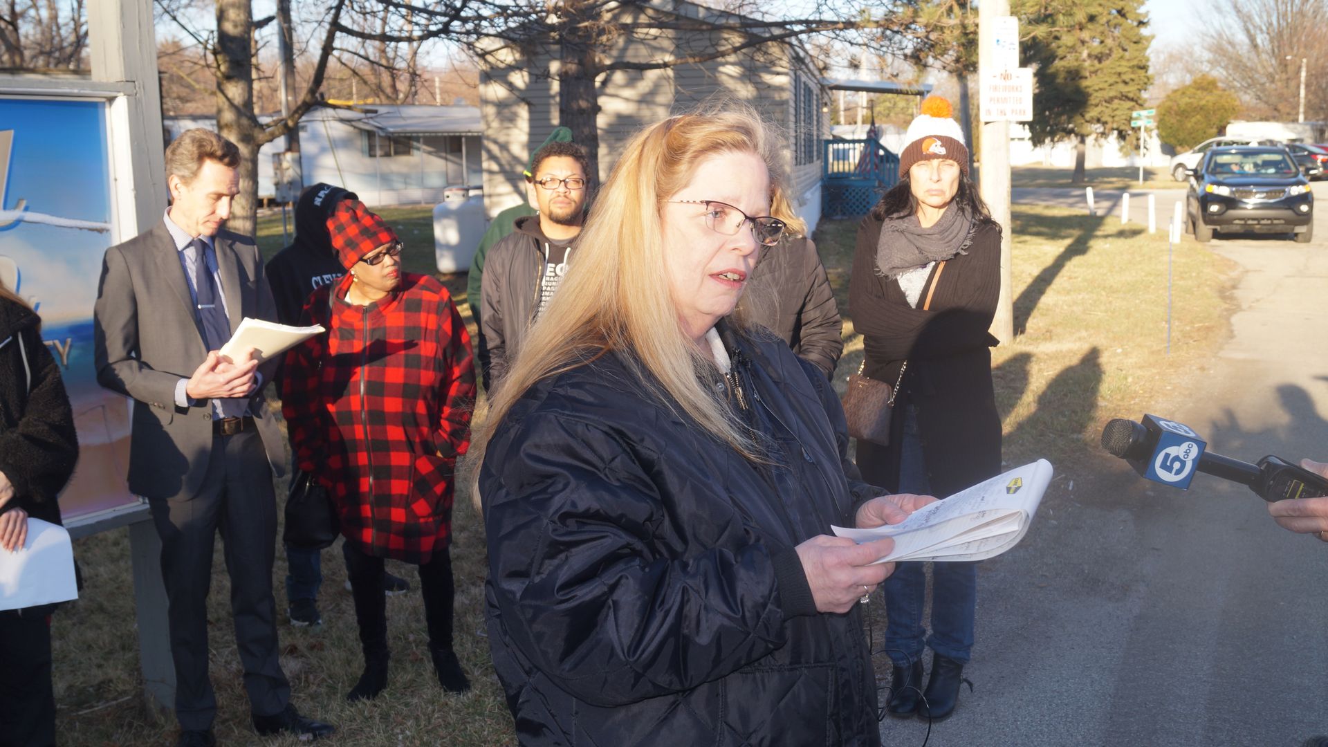 Blonde woman in glasses and black jacket reads from a notepad, addressing reporters.