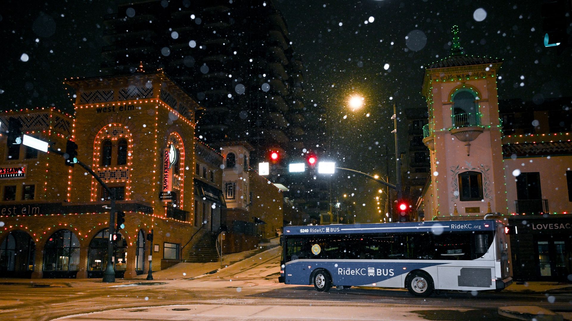 Bus drives across snowy road with nearby building that has windows trimmed with Christmas lights