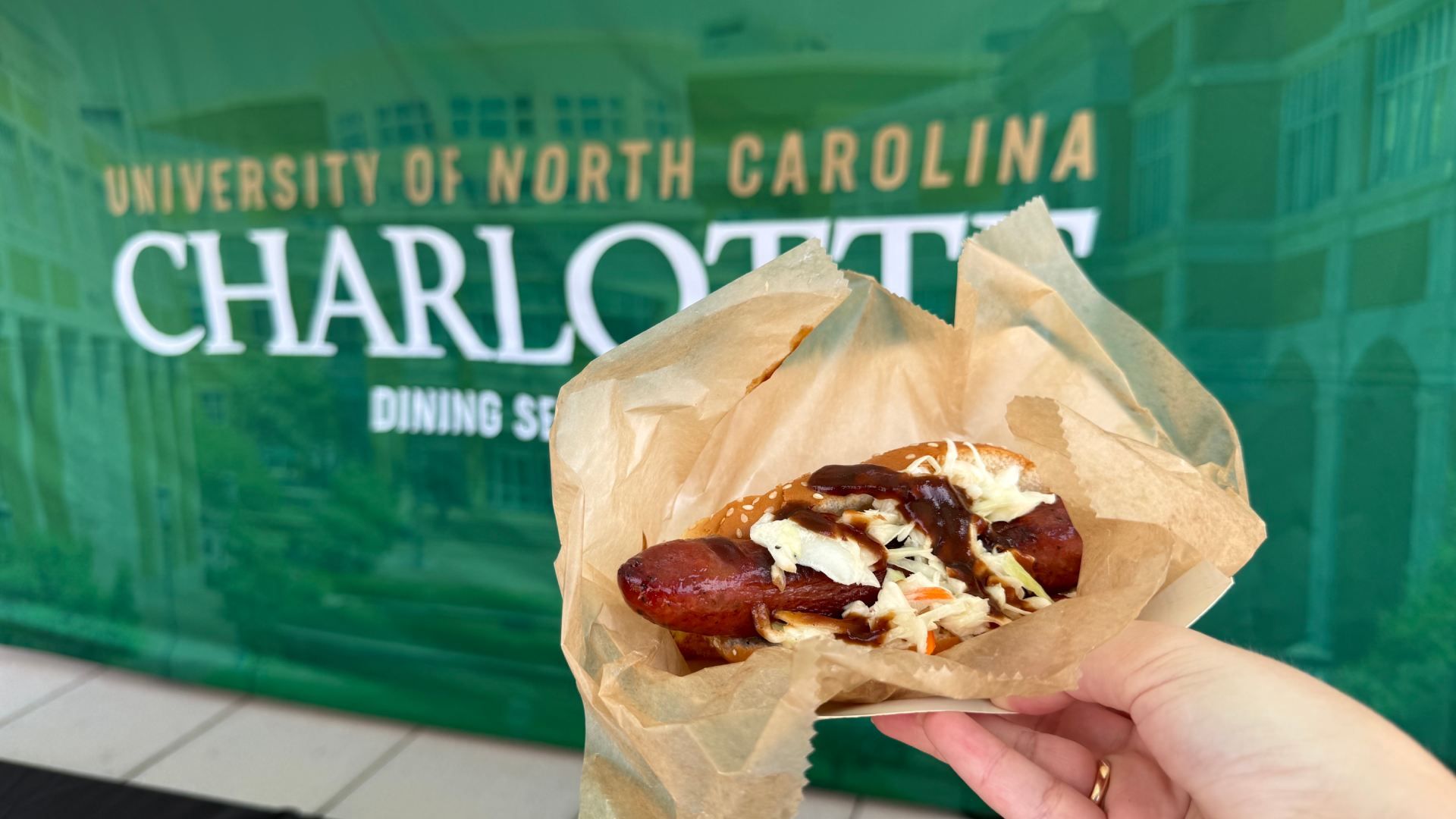 A hand holds a hot dog with sauce and slaw in a crumpled brown paper wrapper, in front of a green storefront window that reads University of North Carolina Charlotte.