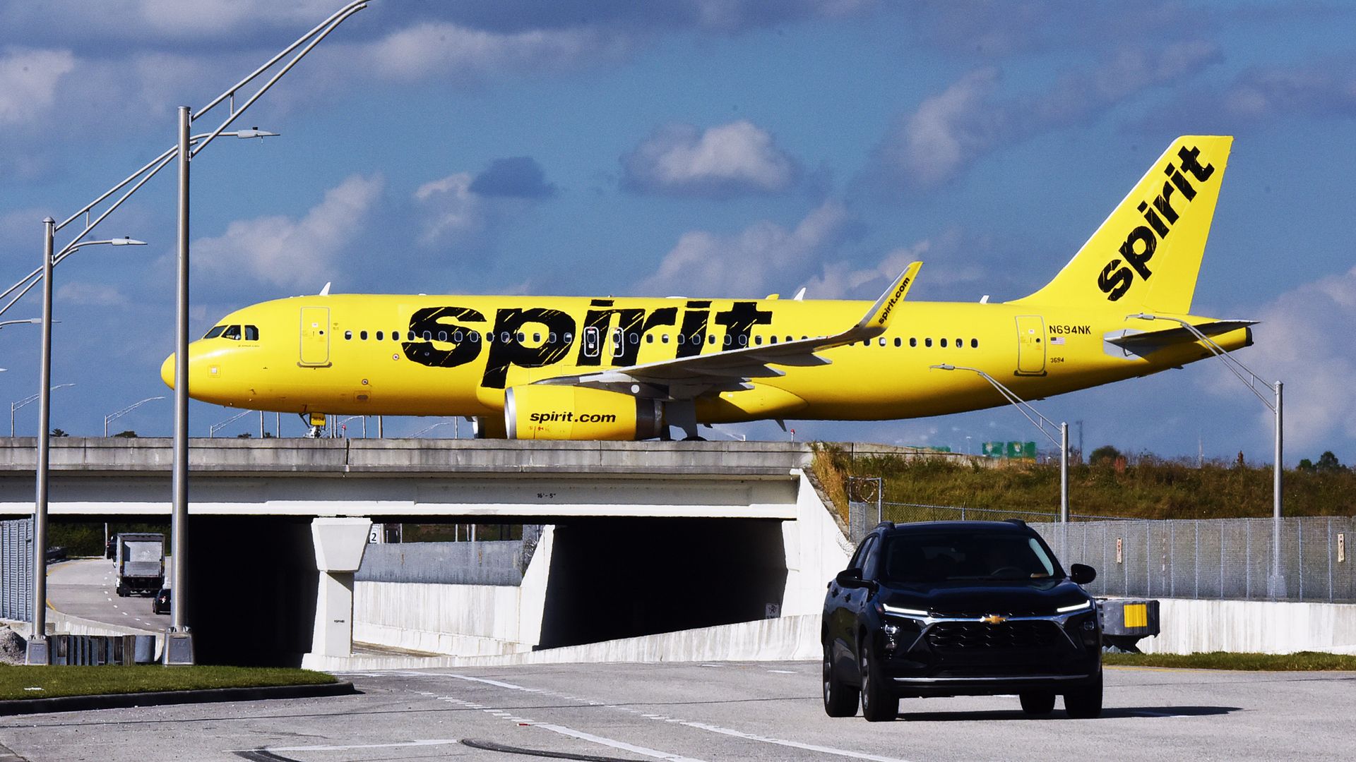Bright yellow Spirit Airlines airplane taxiing on runway overpass above a road with a black car driving below under a partly cloudy sky.