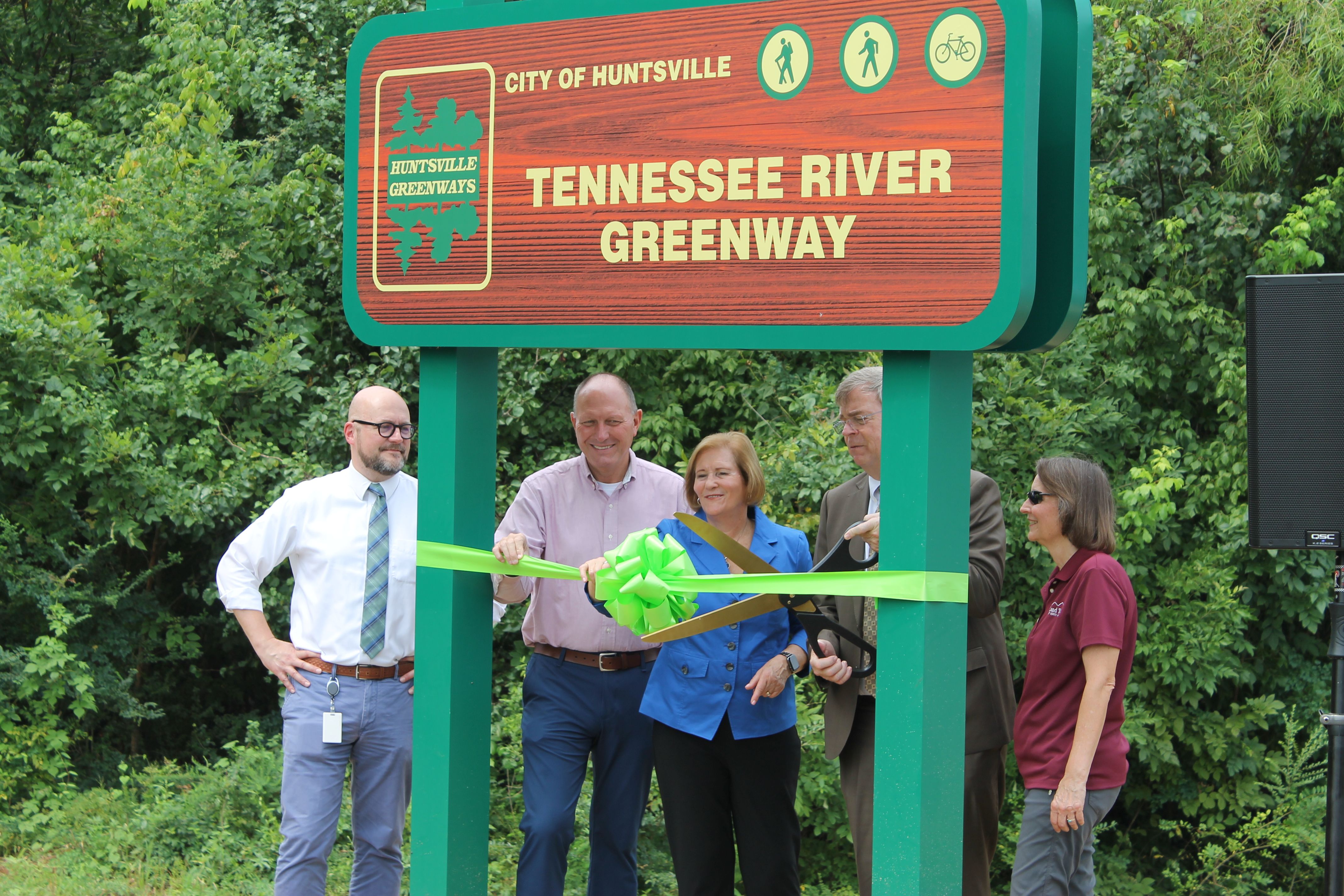 Several professionally dressed people cut a bright green ribbon in front of a large Huntsville sign reading "Tennessee River Greenway," surrounded by green trees.