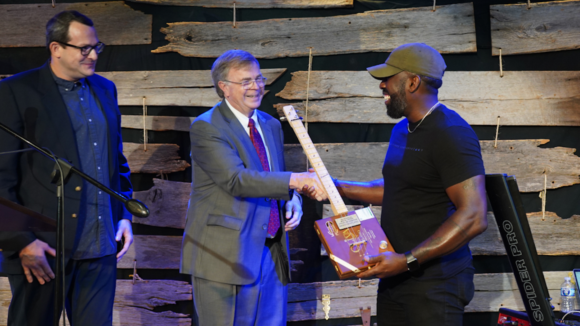 Two men shaking hands on stage with a wooden guitar; one in a gray suit and one in a black t-shirt and green cap, another man in a dark suit watches smiling, rustic wood backdrop.