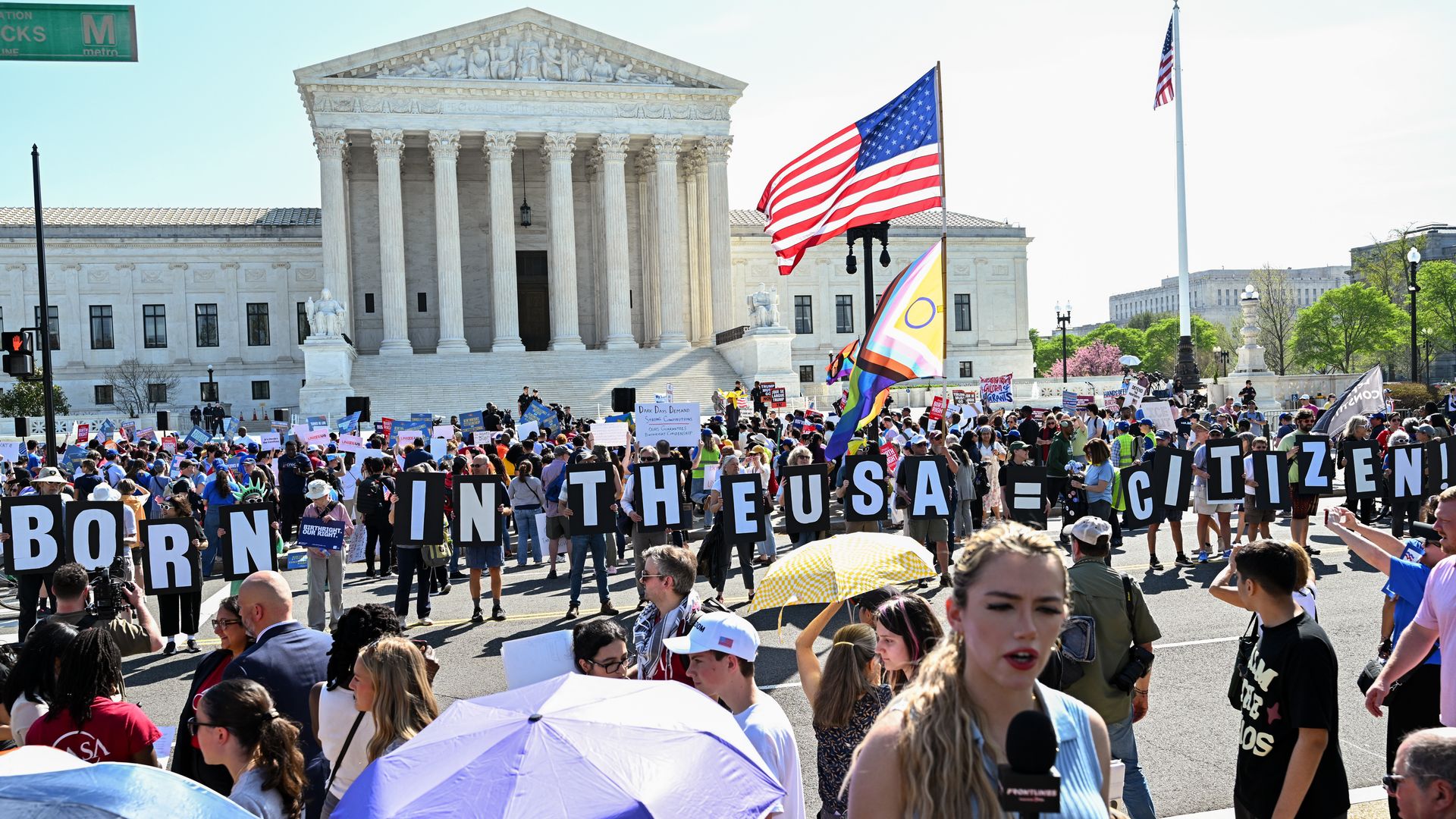 Protesters fill the steps of the U.S. Supreme Court with signs reading "BORN IN THE USA = CITIZEN"; a reporter with a microphone stands in the foreground, American and rainbow flags visible.