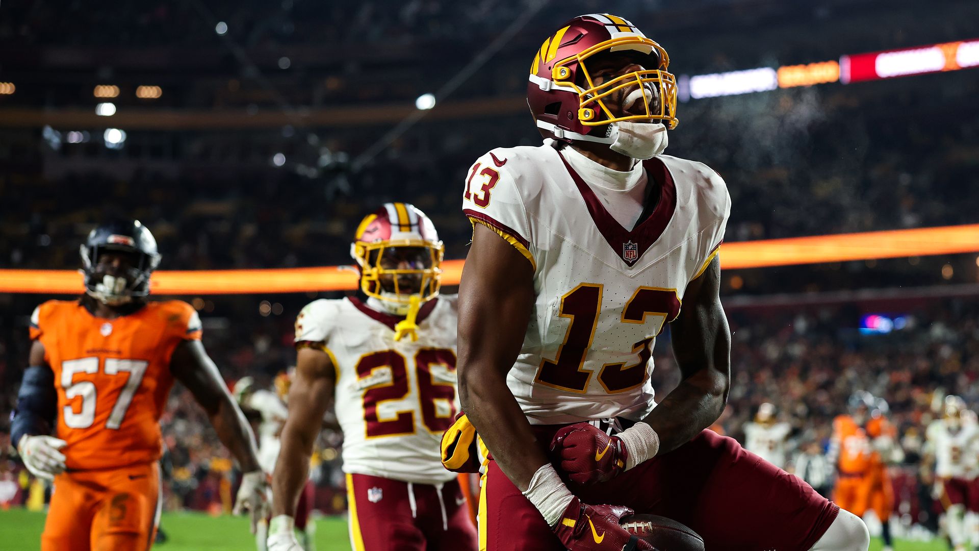 Football player in white and burgundy uniform, number 13, celebrating with a football in hand on a green field with stadium lights and players in orange and white in the background.