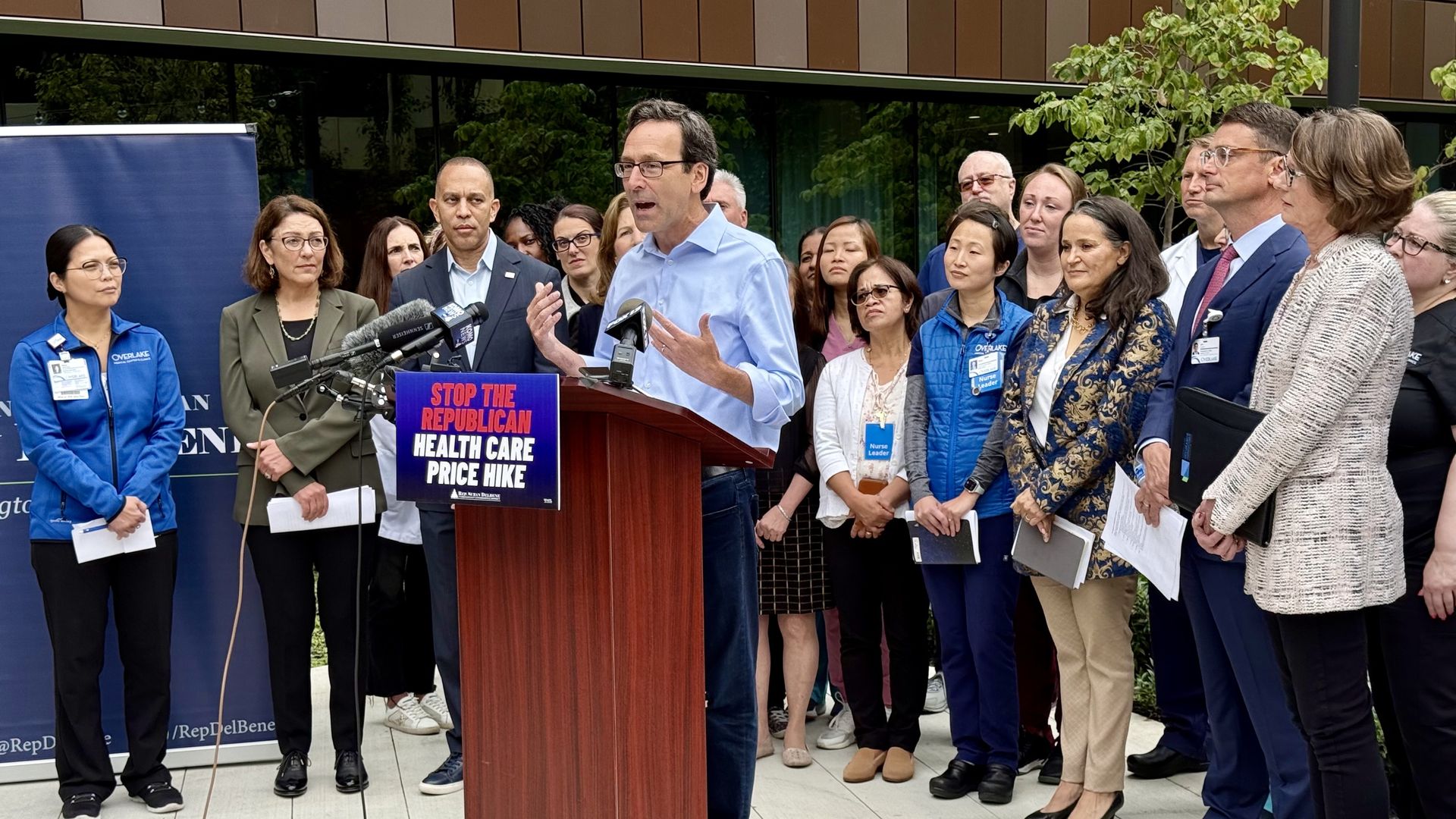 Man speaking at podium with sign "Stop the Republican Health Care Price Hike," surrounded by diverse group of people, some wearing medical badges and uniforms, outside a modern building.