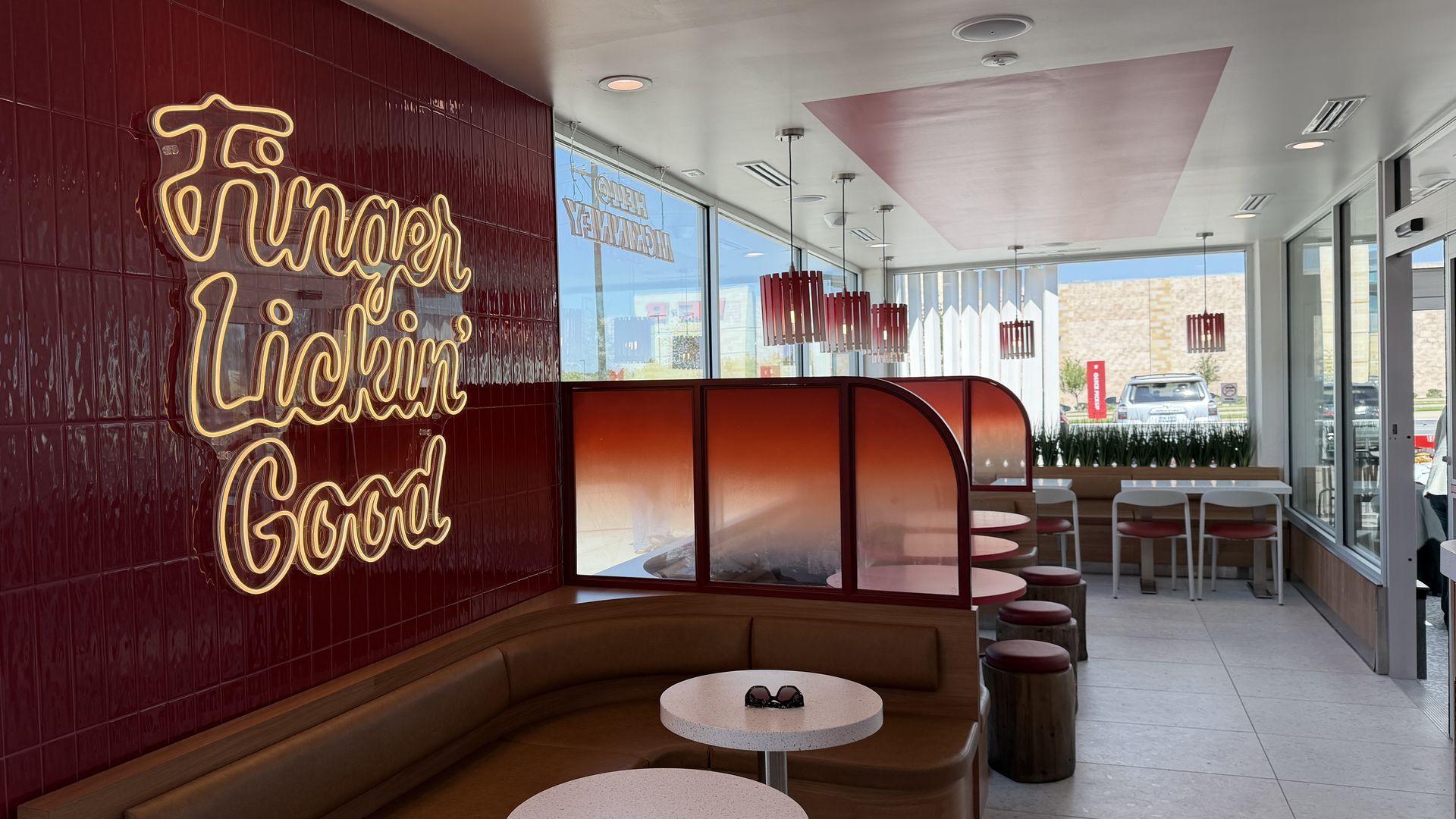Inside a retro cafe with maroon tiled wall and a large yellow neon cursive sign. Brown curved booths, white round tables, red pendant lights, and large windows facing a parking lot.