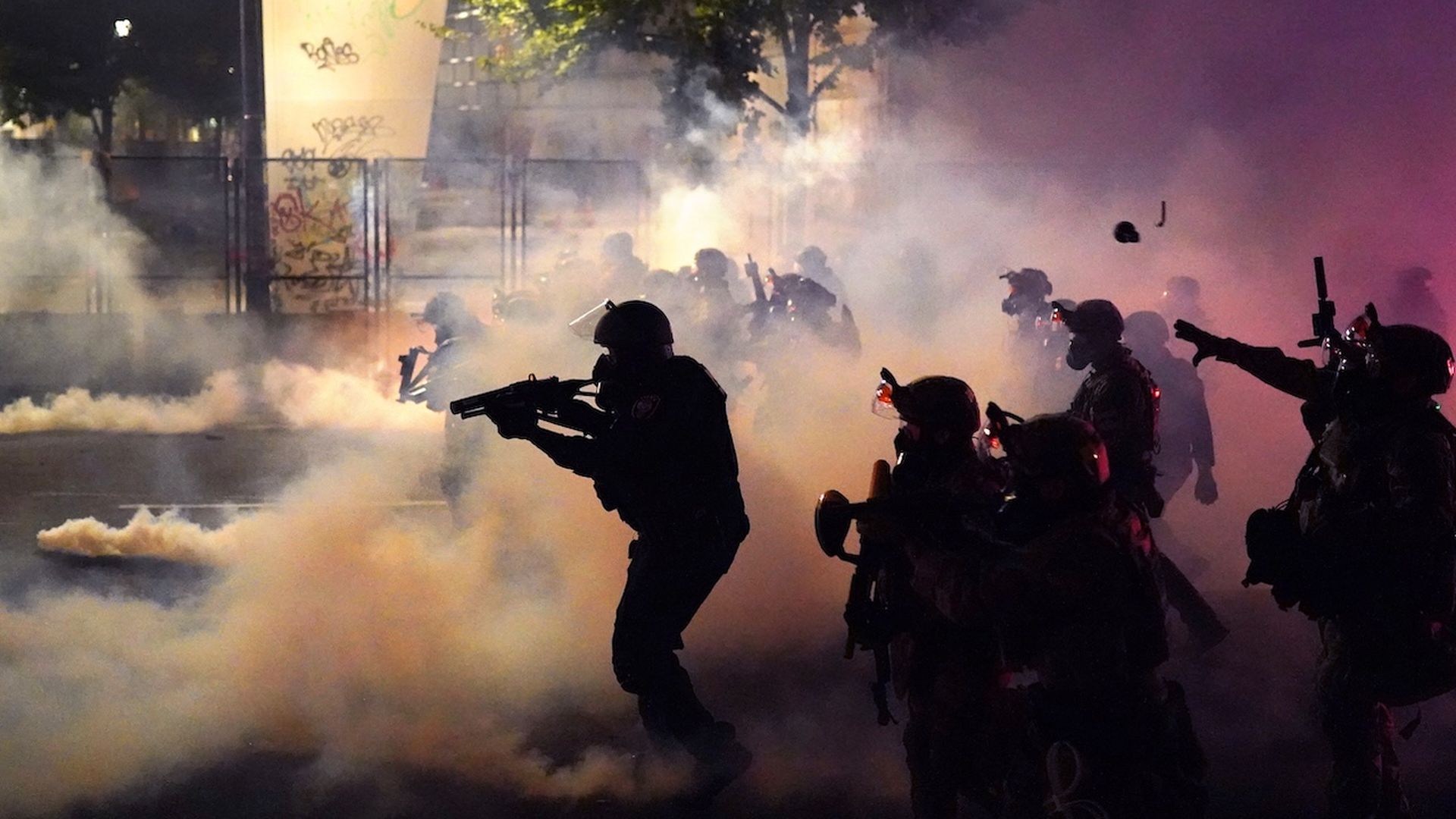 A line of police officers stand in a formation during the night, where thick clouds of tear gas waft. One officer throws a projectile.