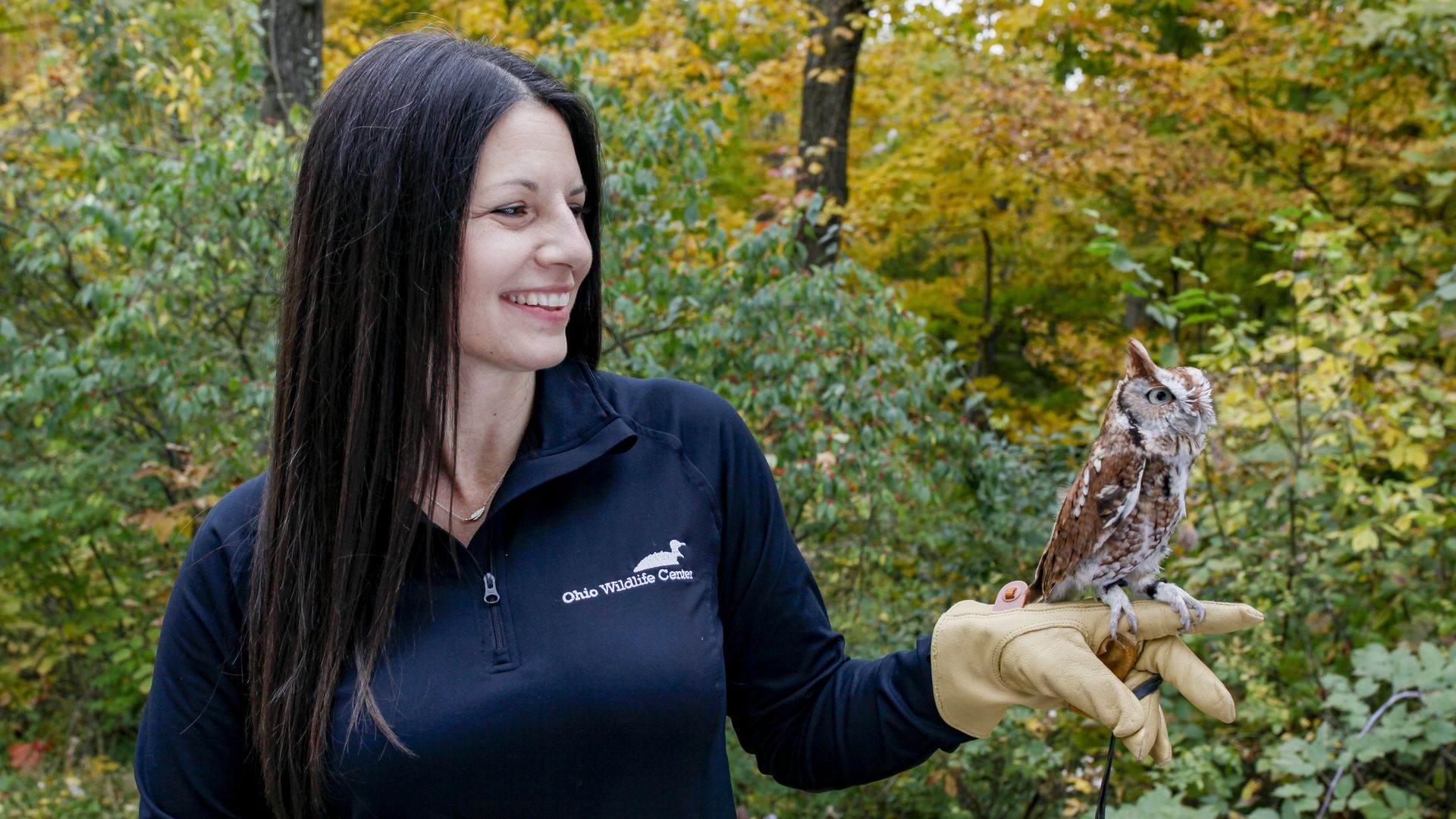 Ohio Wildlife Center executive director Lolita Haverlock holds a small owl on her finger in a nature setting.