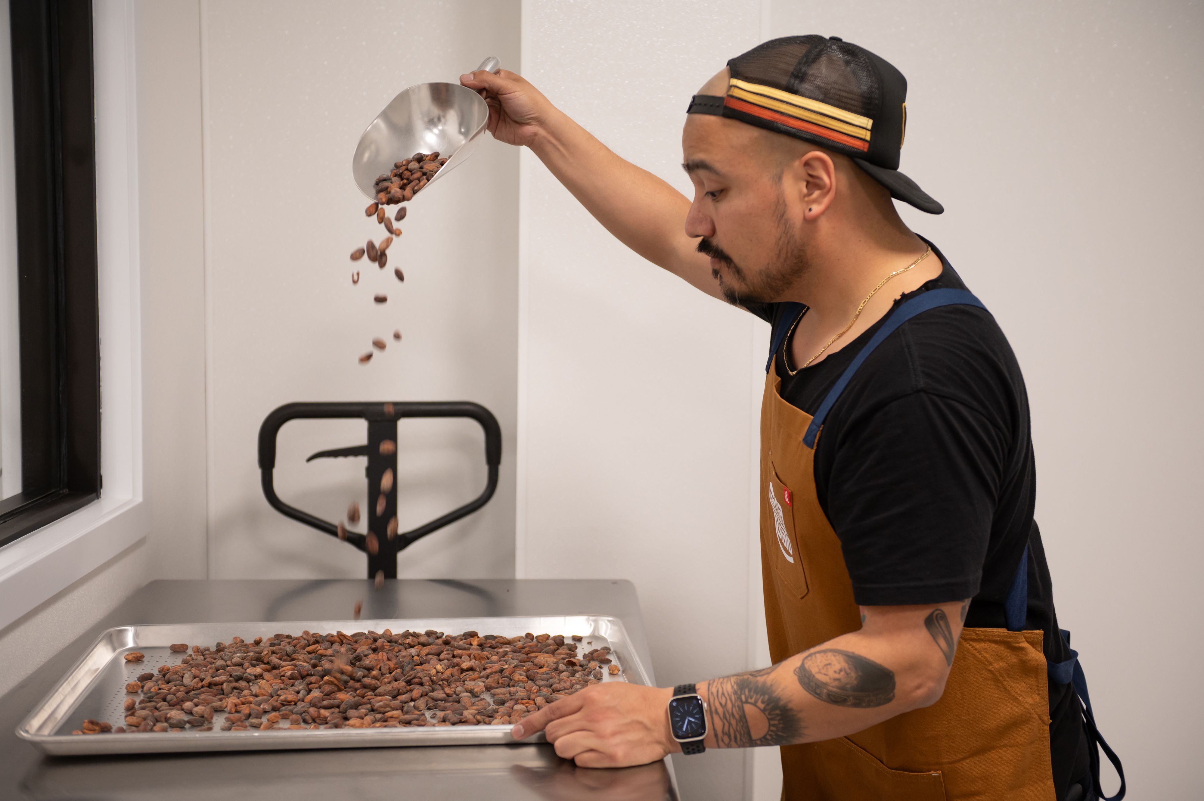 Aaron Lindstrom scoops beans that fall through the air at Cocoa Legato, wearing an apron and hat and black tee shirt.
