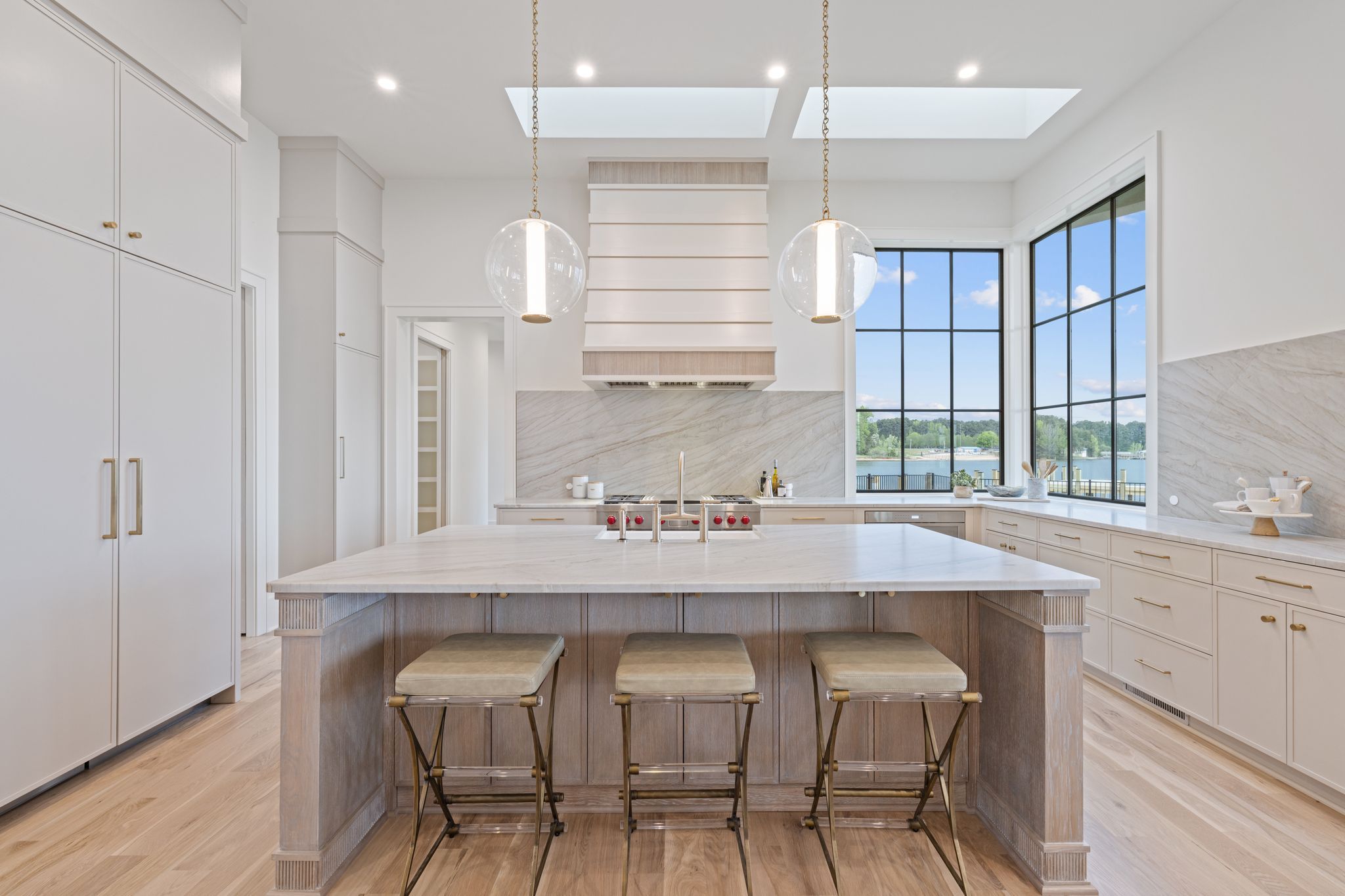 Bright modern kitchen with white and light wood cabinets, marble countertops, three stools at island, two globe pendant lights, large black-framed windows showing a water view.