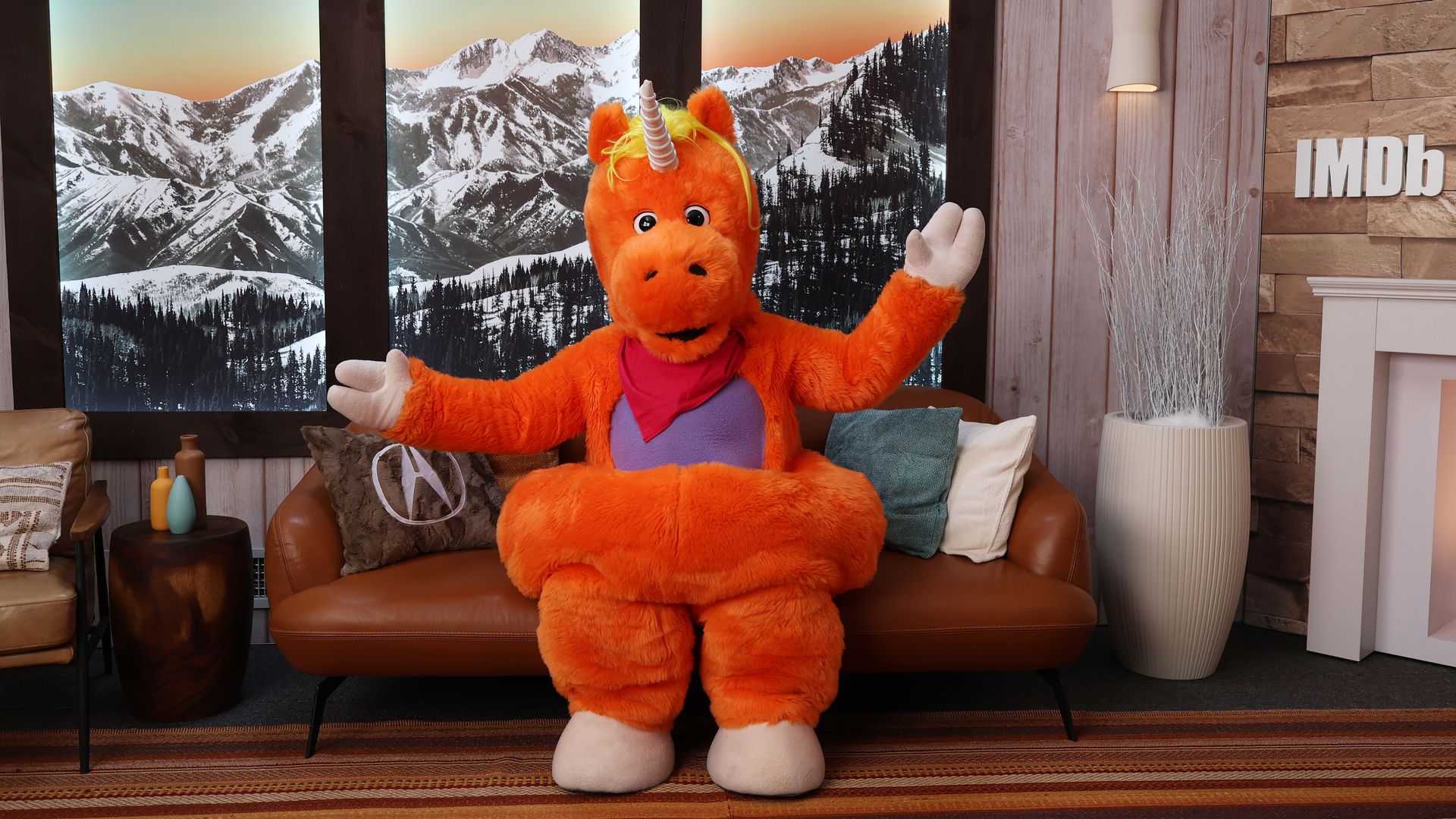Bright orange plush unicorn with a horn and yellow mane sits on a brown leather sofa, arms raised, in front of a snowy mountain mural at sunset; IMDb letters on the wall to the right.