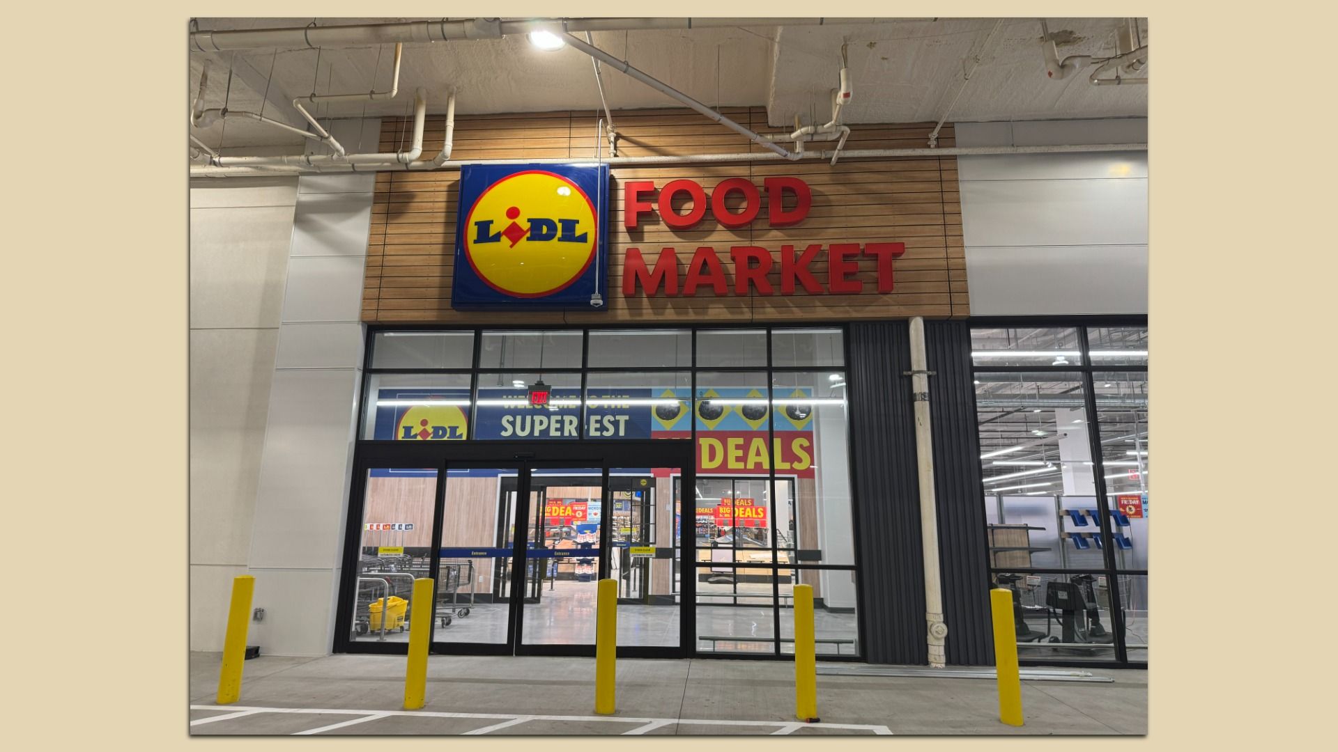 Exterior of a Lidl Food Market store with a bright yellow and blue Lidl logo and red "Food Market" letters on wood paneling above glass entrance doors. Yellow bollards line the sidewalk.