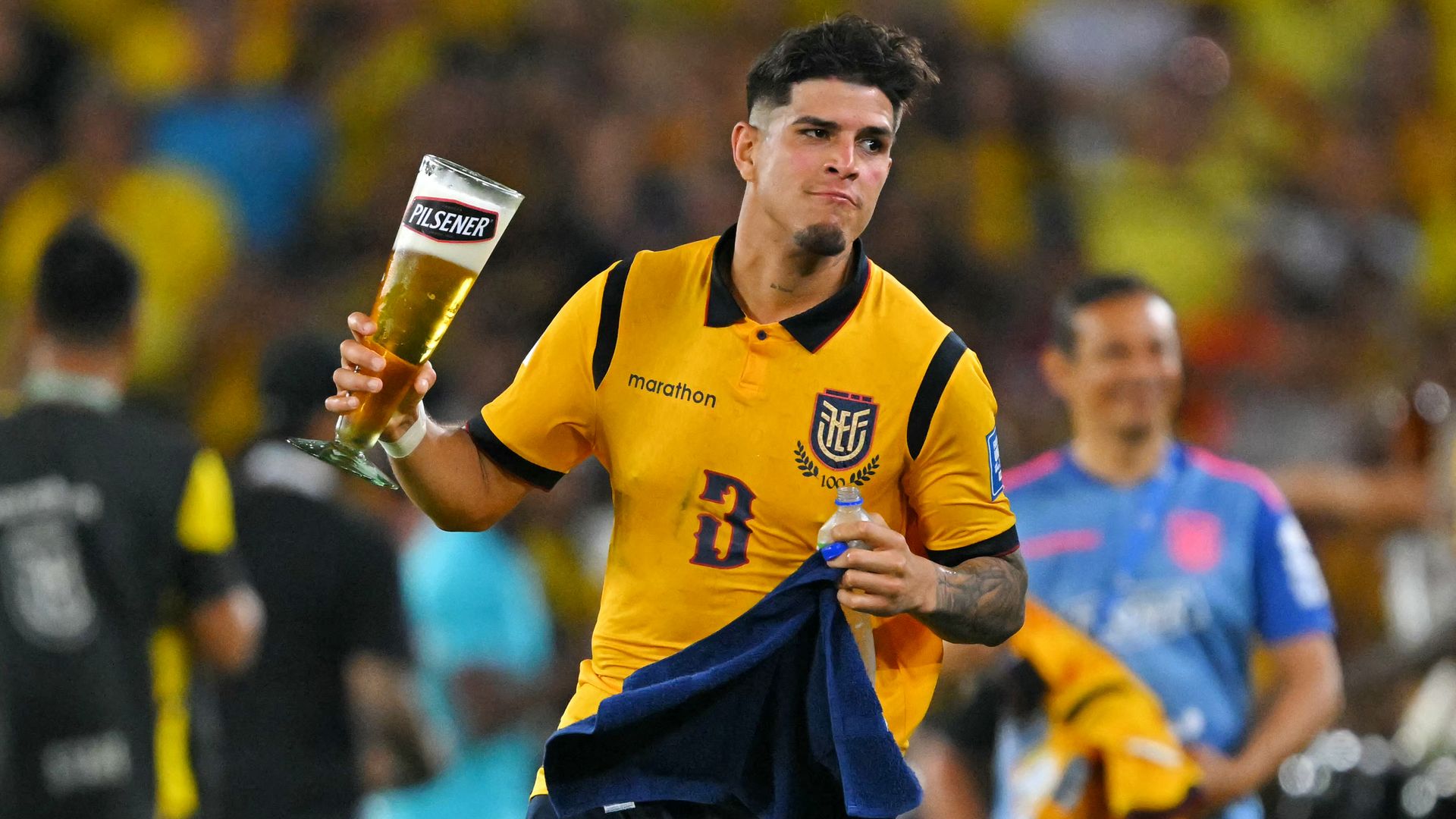 A soccer player in a yellow jersey holds a tall glass labeled PILSENER in one hand and a water bottle in the other, with a blurred yellow-clad crowd behind him.
