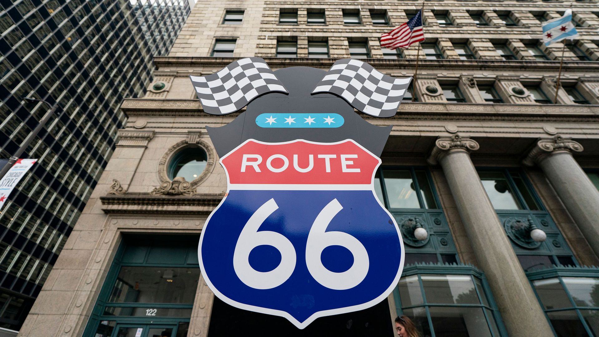 Large Route 66 shield sign with checkered racing flags on top, set against tall buildings displaying American and Chicago flags.