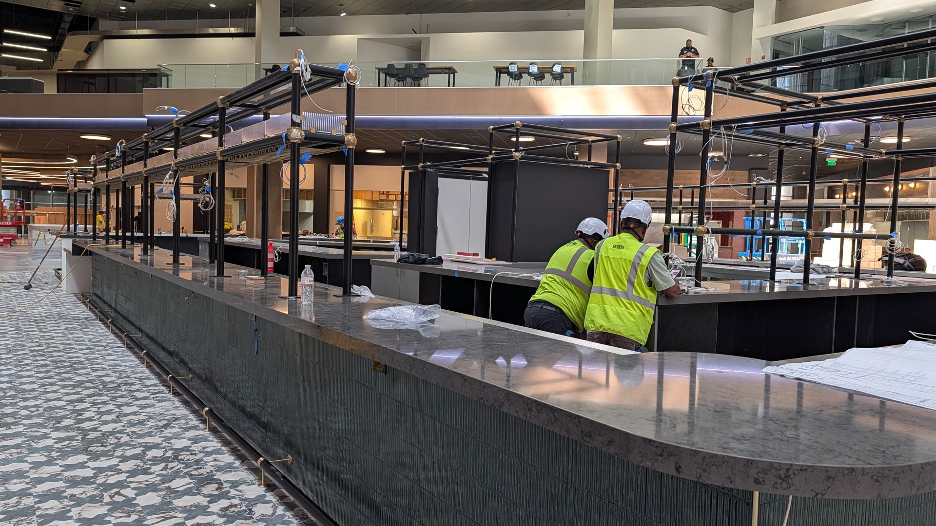 Indoor construction site with a long curved black marble counter beneath a metal framework and hanging wires. Two workers in high-visibility vests and hard hats work; upper level has a glass railing.