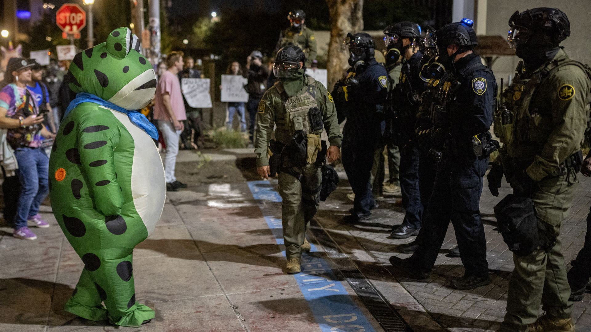 Person in green frog costume with blue scarf faces line of police and armored officers at night, with onlookers and protest signs in the background on city sidewalk.