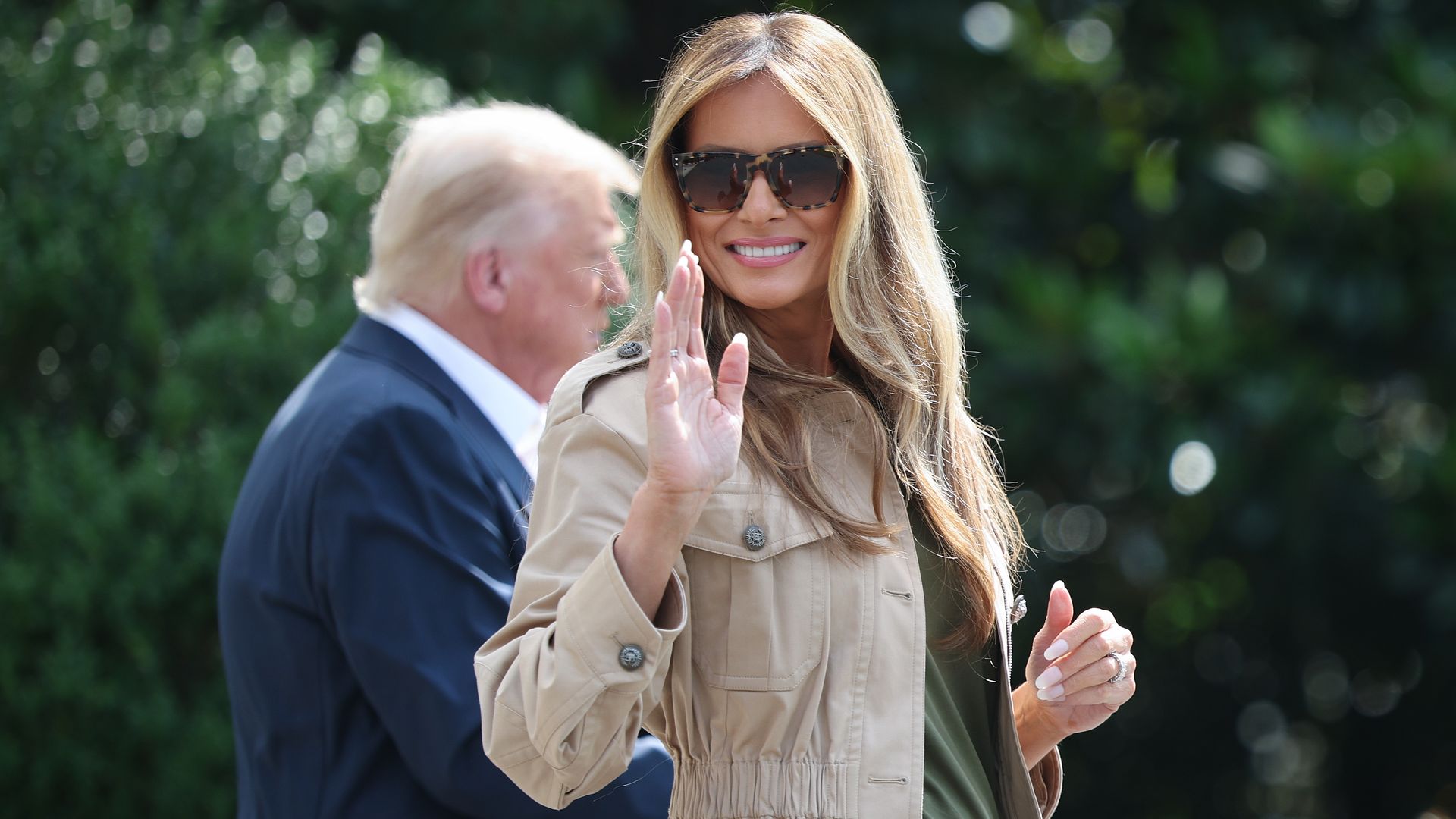 Smiling woman with long blonde hair wearing sunglasses, a beige jacket, and green top, waving with one hand; blurred man in navy suit walking behind her outdoors with greenery.