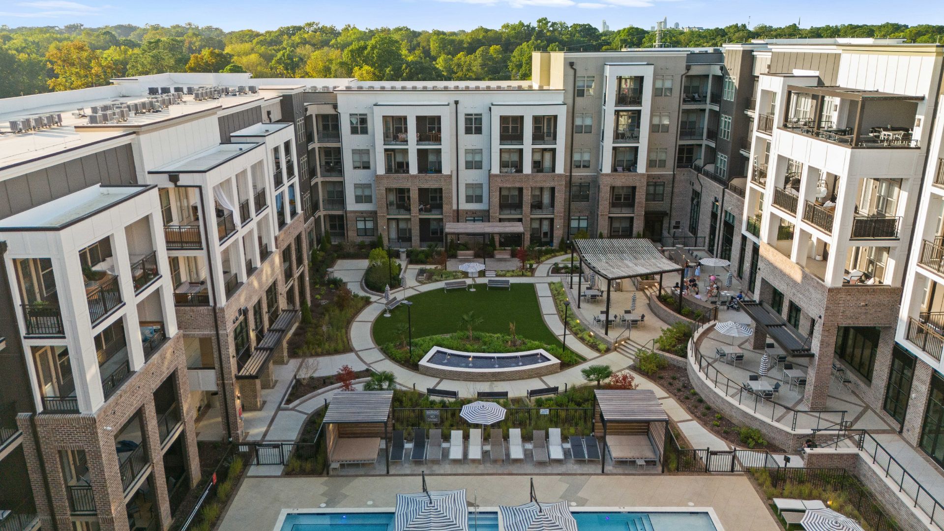 Aerial view of a modern apartment complex courtyard: white and beige buildings surround a green lawn with a circular path, benches, a pergola, striped umbrellas and a pool at the bottom, with trees in the background.