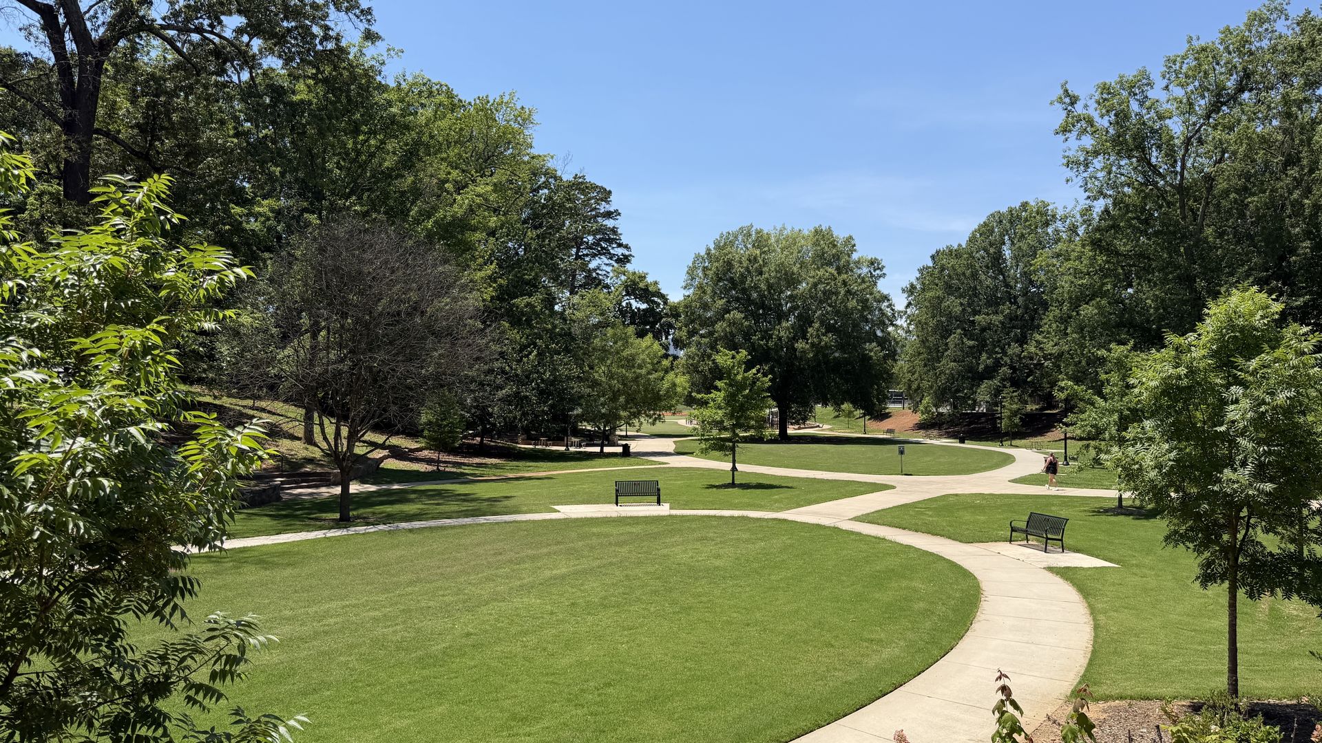Sunny park scene with lush green grass, curving concrete paths, and many trees. A person walks on the right; several benches dot the lawn.