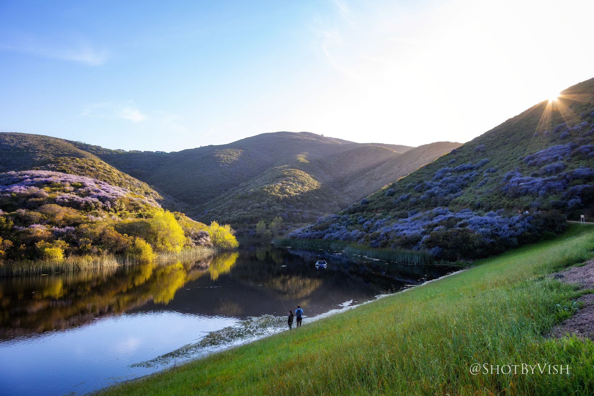 Two people stand at the edge of a lake surrounded by lush green and purple hills.