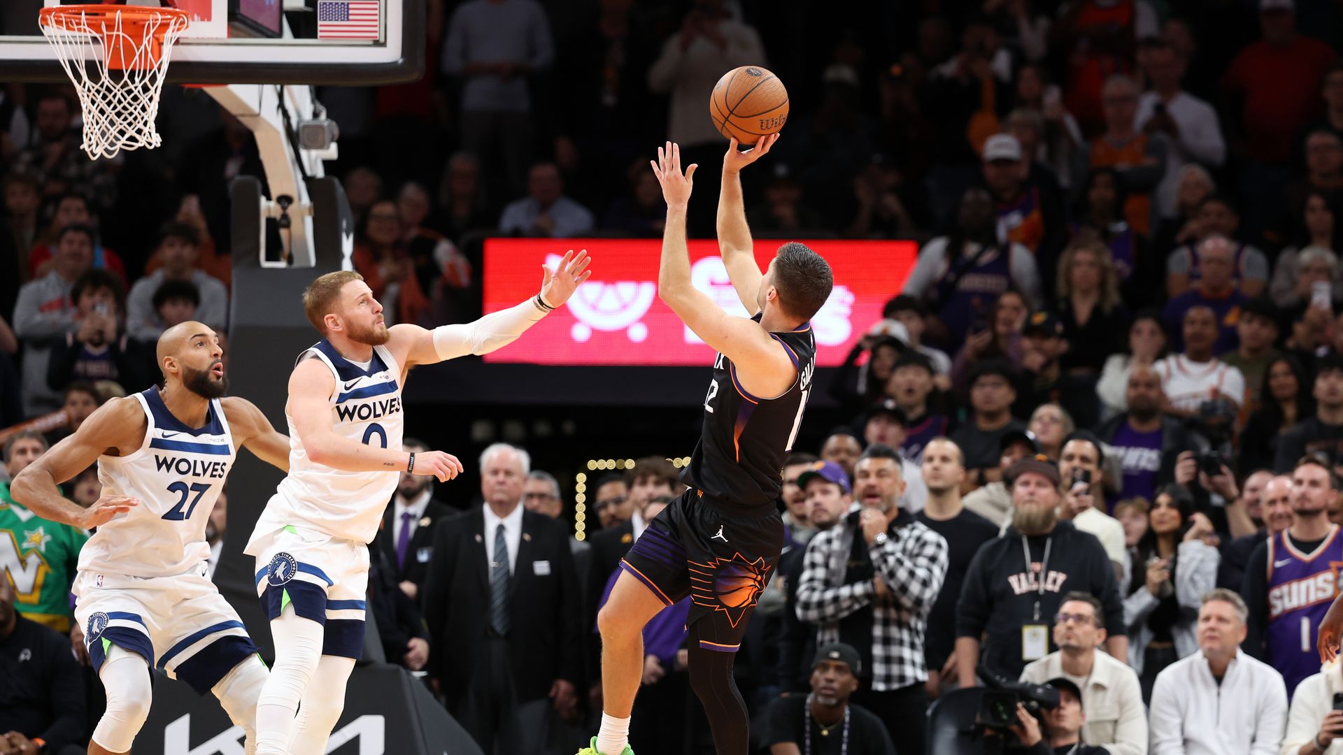 A basketball player in a black uniform shoots the ball mid-air while with two opposing players in white uniforms in front of him. 