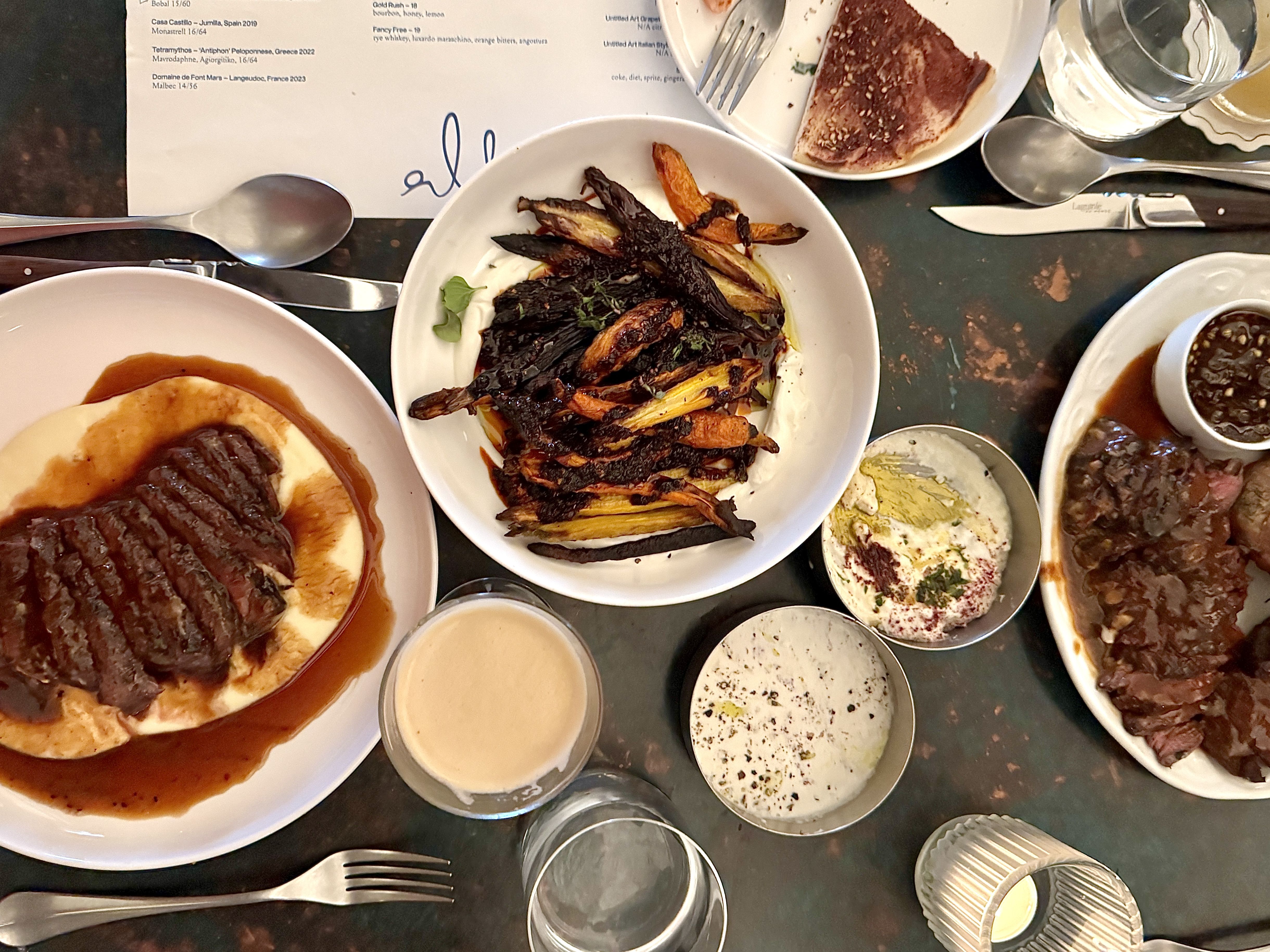 Overhead view of a table with several plates: central bowl of charred roasted vegetables over cream, a beef dish in brown sauce to the left, plus sauces, cutlery, a menu page, and a glass of water.