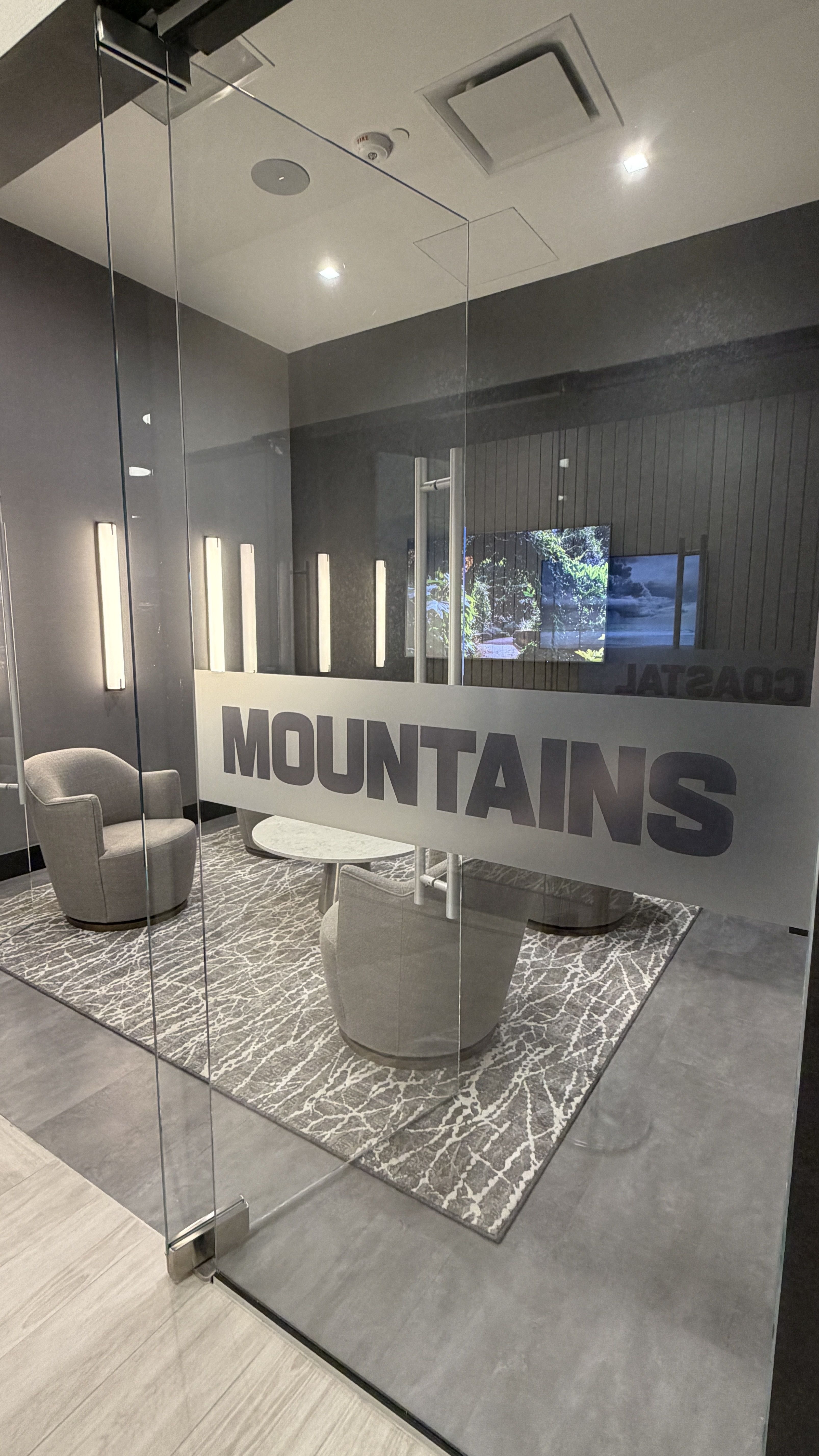 Glass-walled lounge with gray chairs around a white round table on a patterned rug; the frosted glass panel reads "MOUNTAINS" with vertical lights and a TV in the background.
