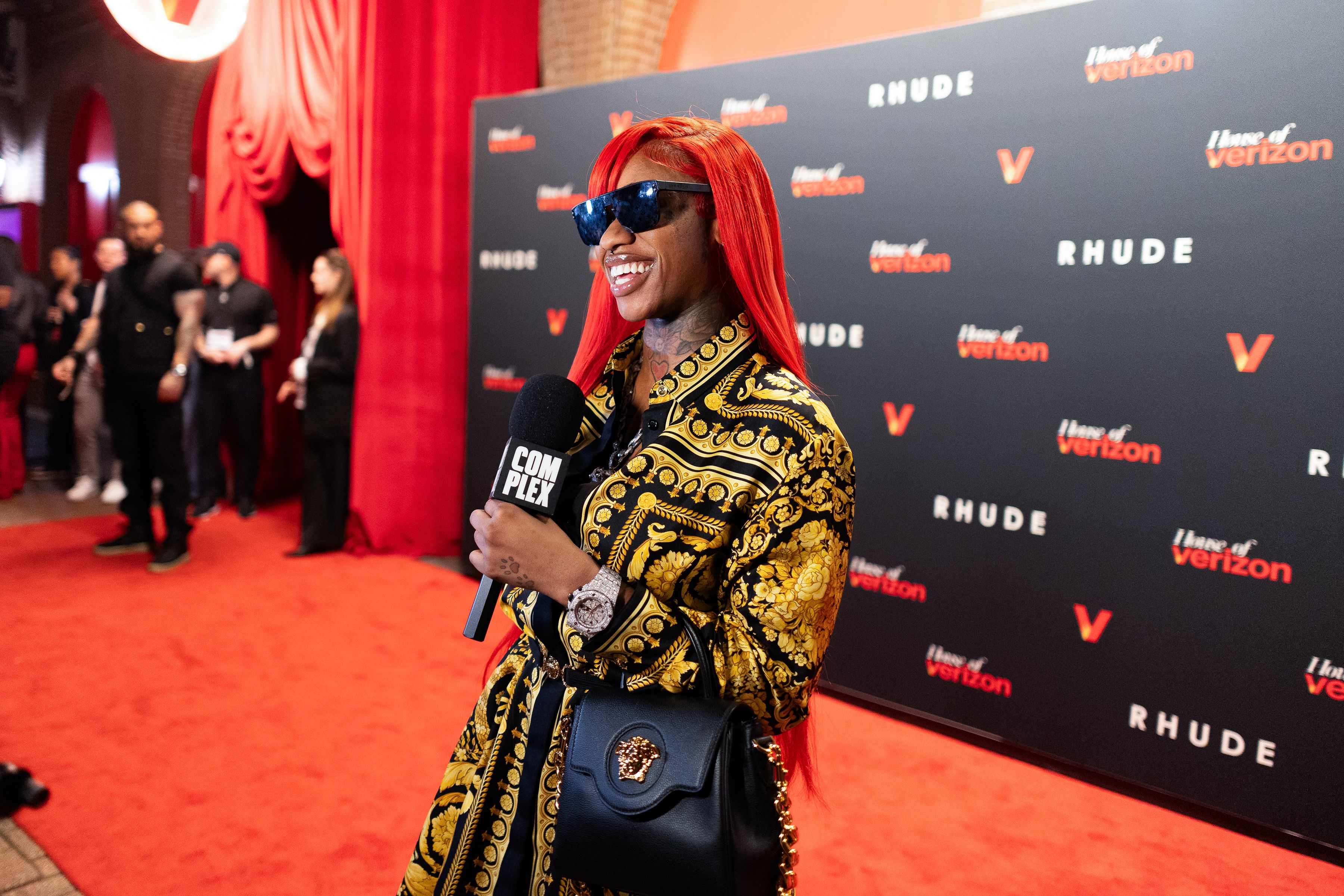 A woman holds a microphone on a red carpet and smiles to someone off-camera.