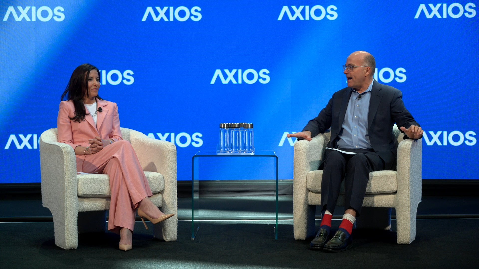 A woman in a pink suit sits next to a man in a blue suit and a light blue shirt in cream-colored chairs on a stage.
