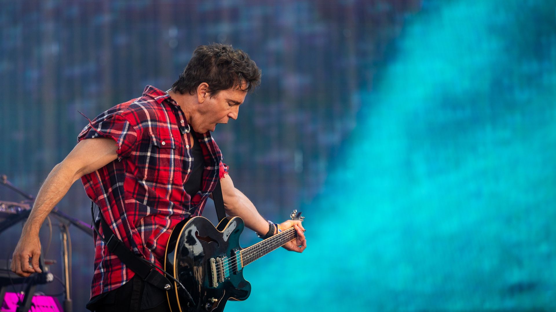 Guitarist wearing a red plaid shirt plays a black electric guitar on stage, with a blue-green, abstract background.