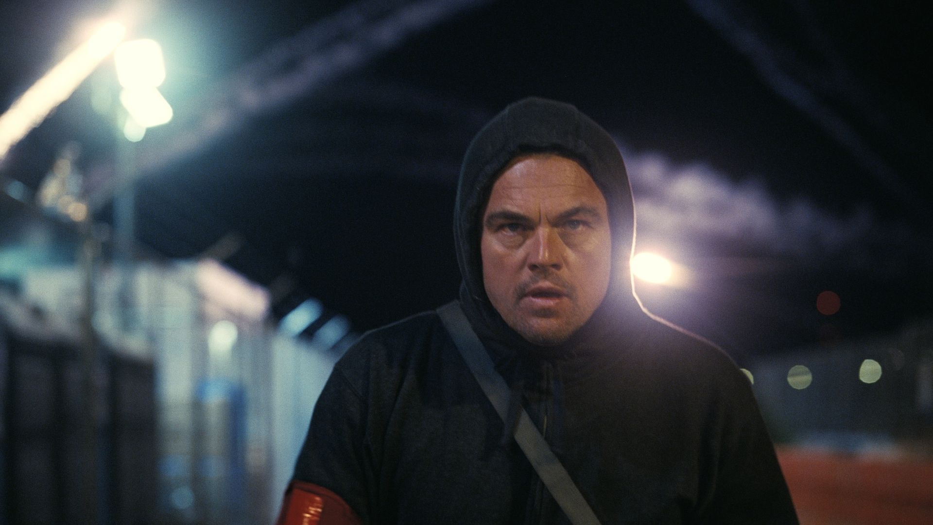 Man with determined expression wearing a black hoodie and red armband, walking at night near a fence with bright lights and smoke trails in the background.