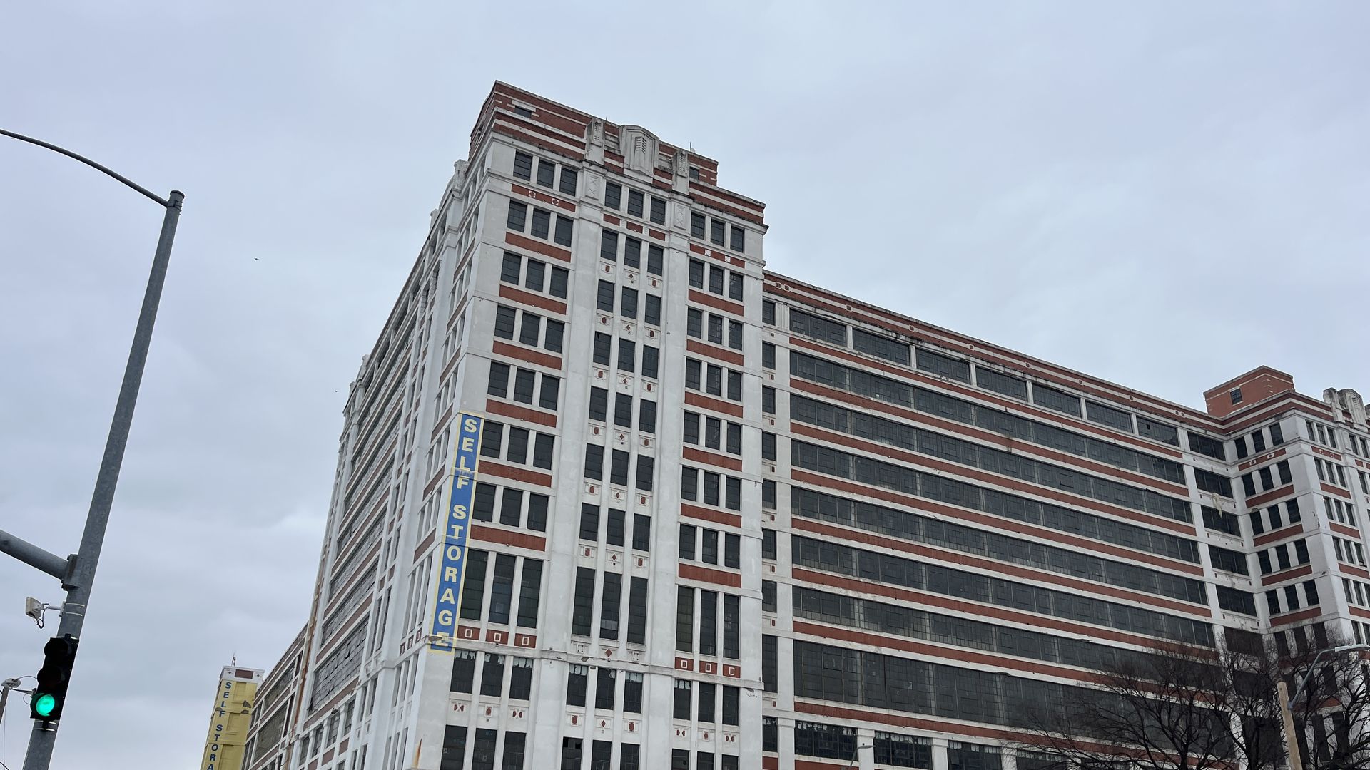 Large multi-story white and red brick building with many windows and vertical "SELF STORAGE" sign on side under cloudy gray sky.