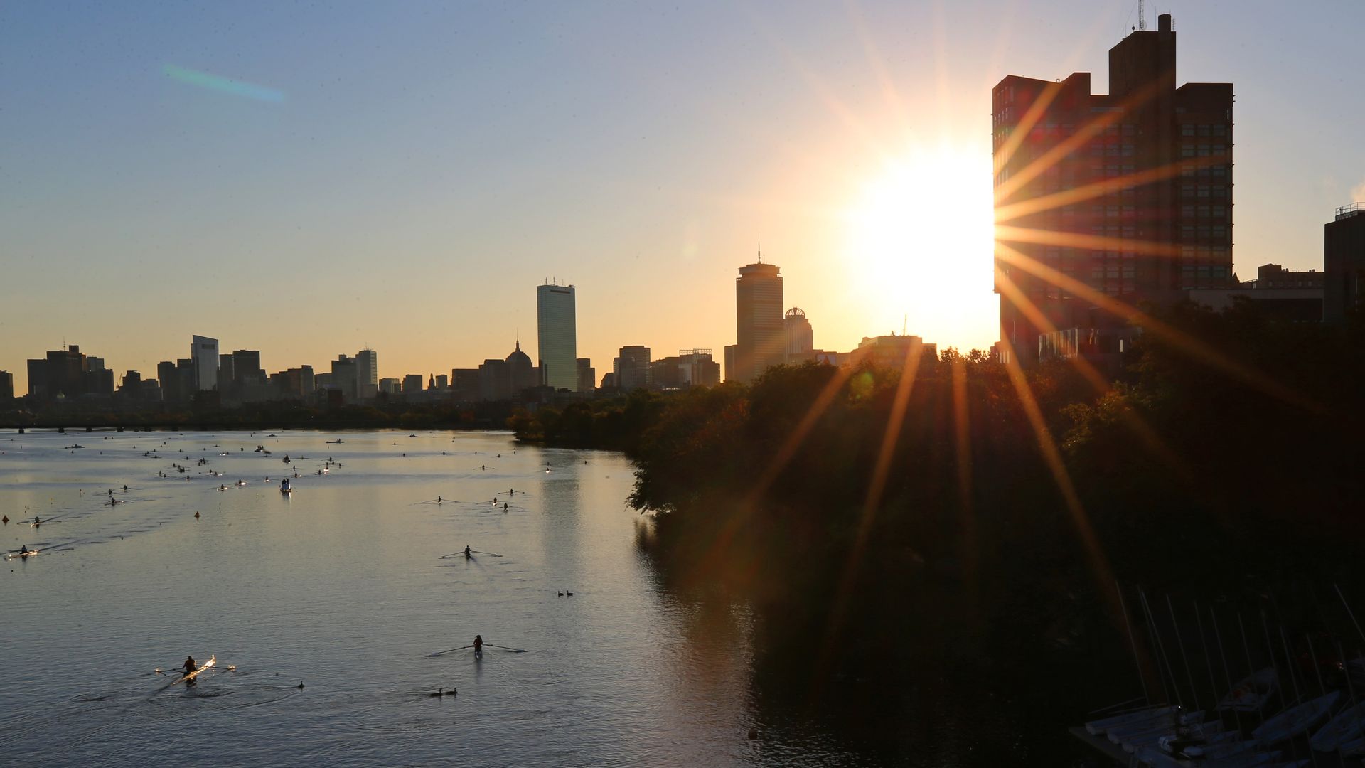 The sun shines over the Boston skyline as rowboats move on the Charles River.