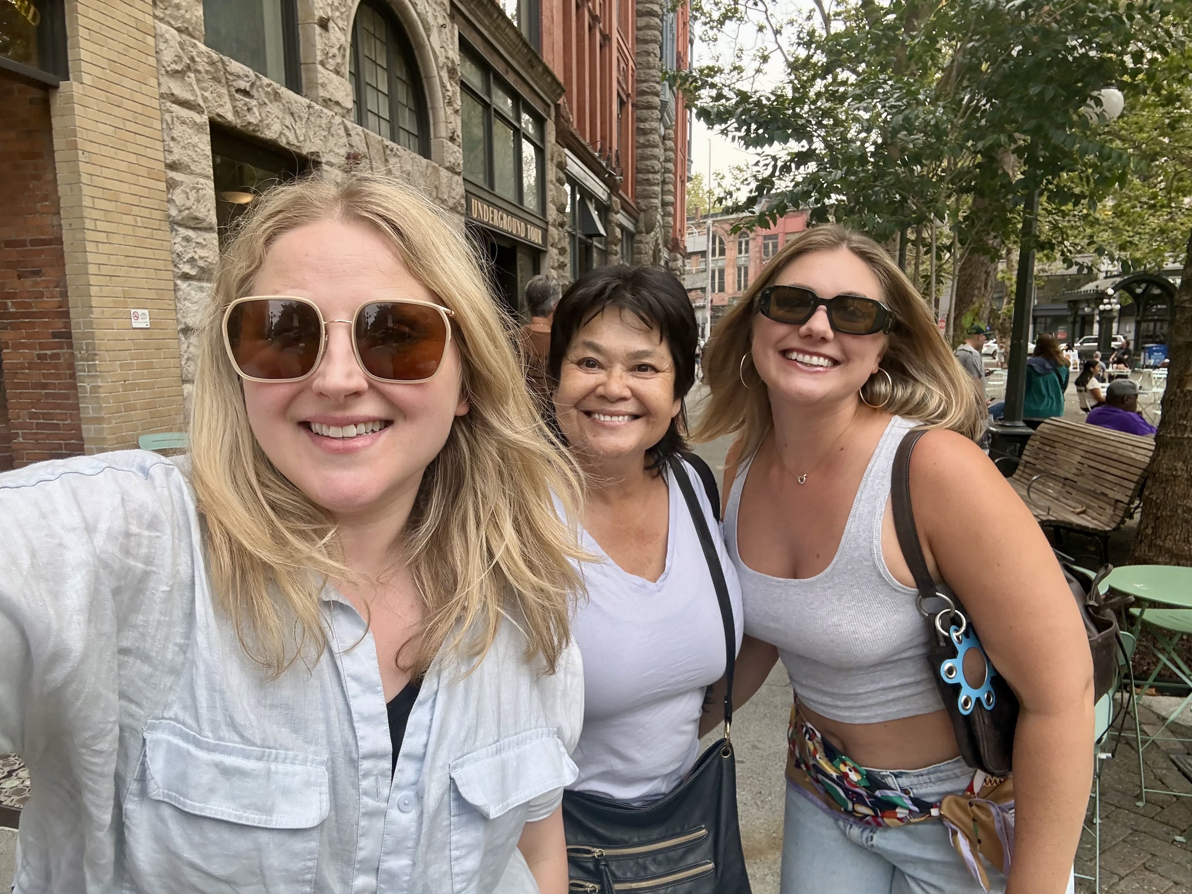 Three smiling women take a selfie on a city sidewalk, with brick and stone buildings and trees in the background; a sign reads "Underground Tour".