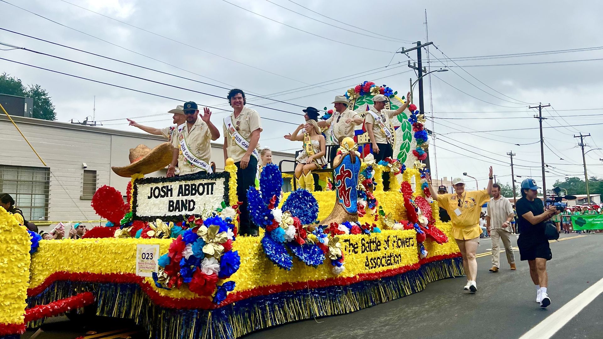 Colorful parade float covered in yellow, blue, and red flowers with fringe along the sides. People in light shirts wave from the float; signs read "Josh Abbott Band" and "Battle of Flowers Association" as spectators line the street.