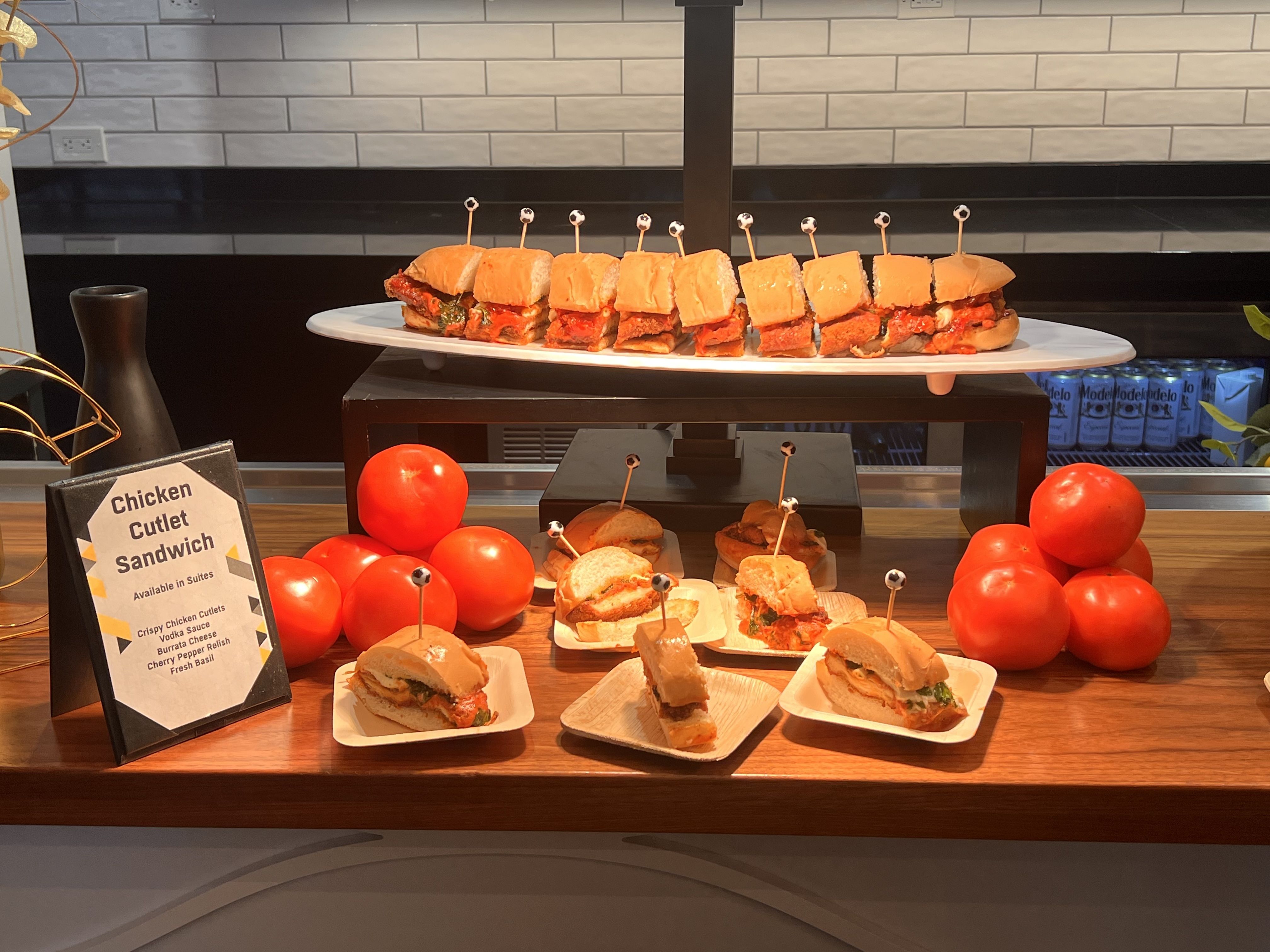 Display of chicken cutlet sandwiches on plates and a white platter, with toothpicks featuring black and white ball tops. Red tomatoes stacked beside a sign describing sandwich ingredients.