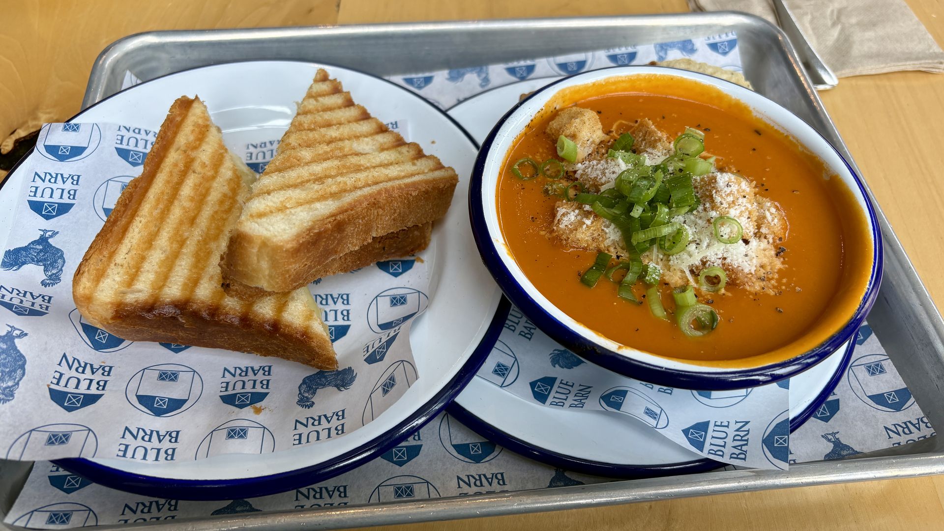 Two triangular grilled cheese sandwiches on a Blue Barn liner, beside a blue-rimmed bowl of orange soup topped with croutons, grated cheese, and chopped green onions on a metal tray.